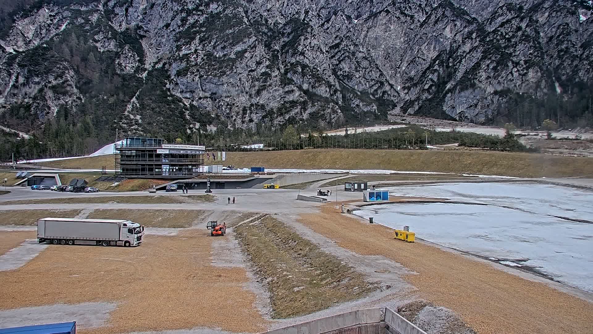 A snowy winter scene reveals a modern multi-story building surrounded by parked cars and an excavator, two small helicopters on a snow-dusted tarmac, and what appears to be a bobsled track, all set against a backdrop of snow-covered mountains partially obscured by thick, gray overcast clouds.