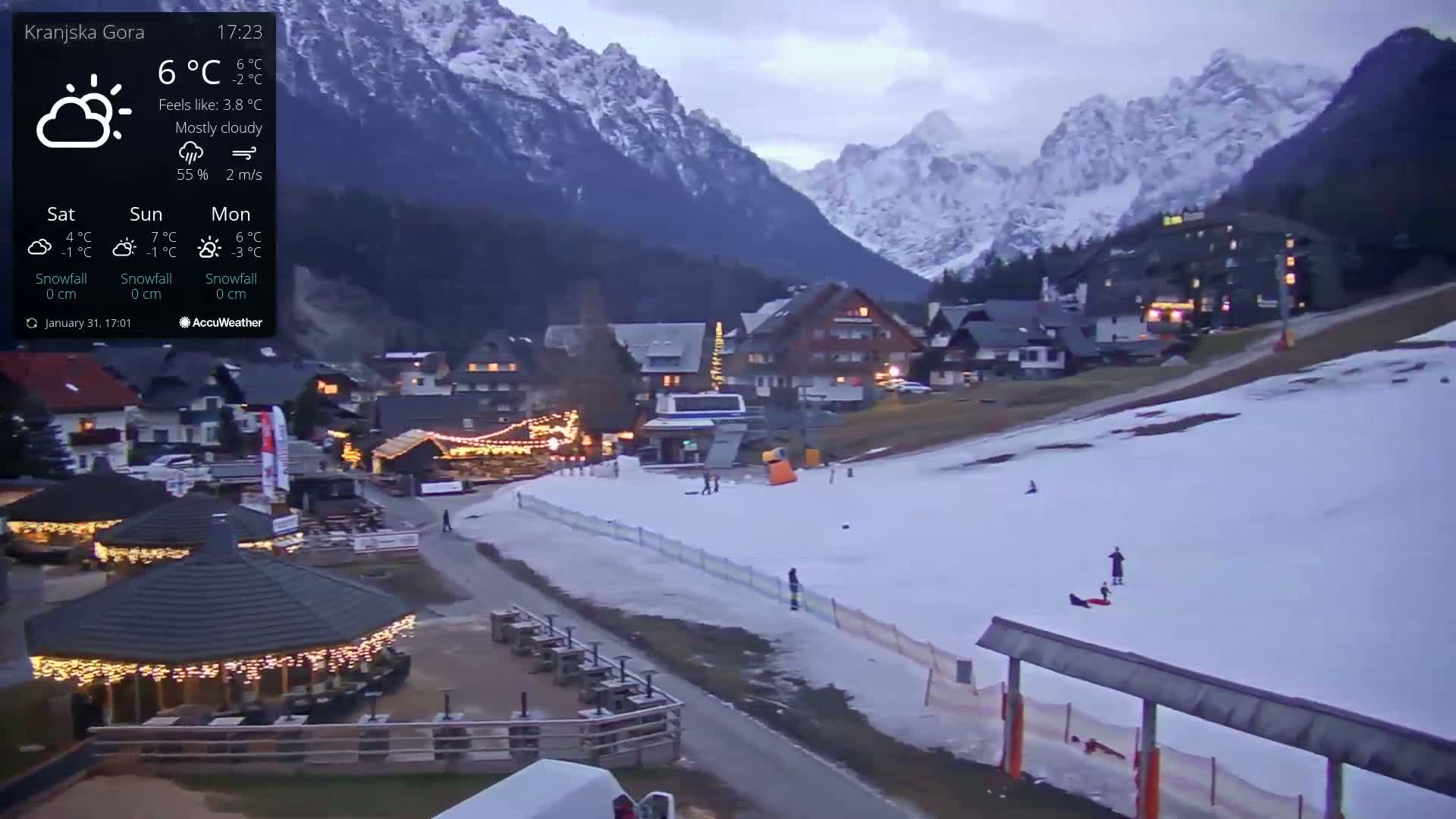 A mostly cloudy evening scene shows a snow-covered slope with a few people and a small village nestled at the base of snow-capped mountains.