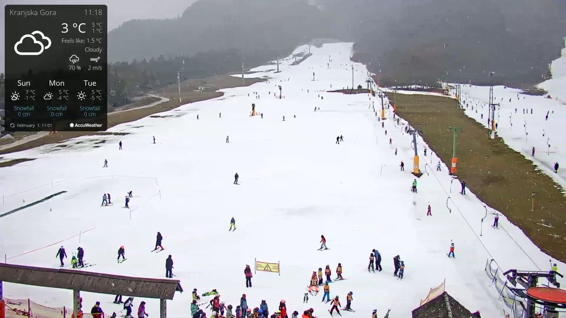 A mostly snow-covered ski slope with numerous skiers and snowboarders, under cloudy conditions.