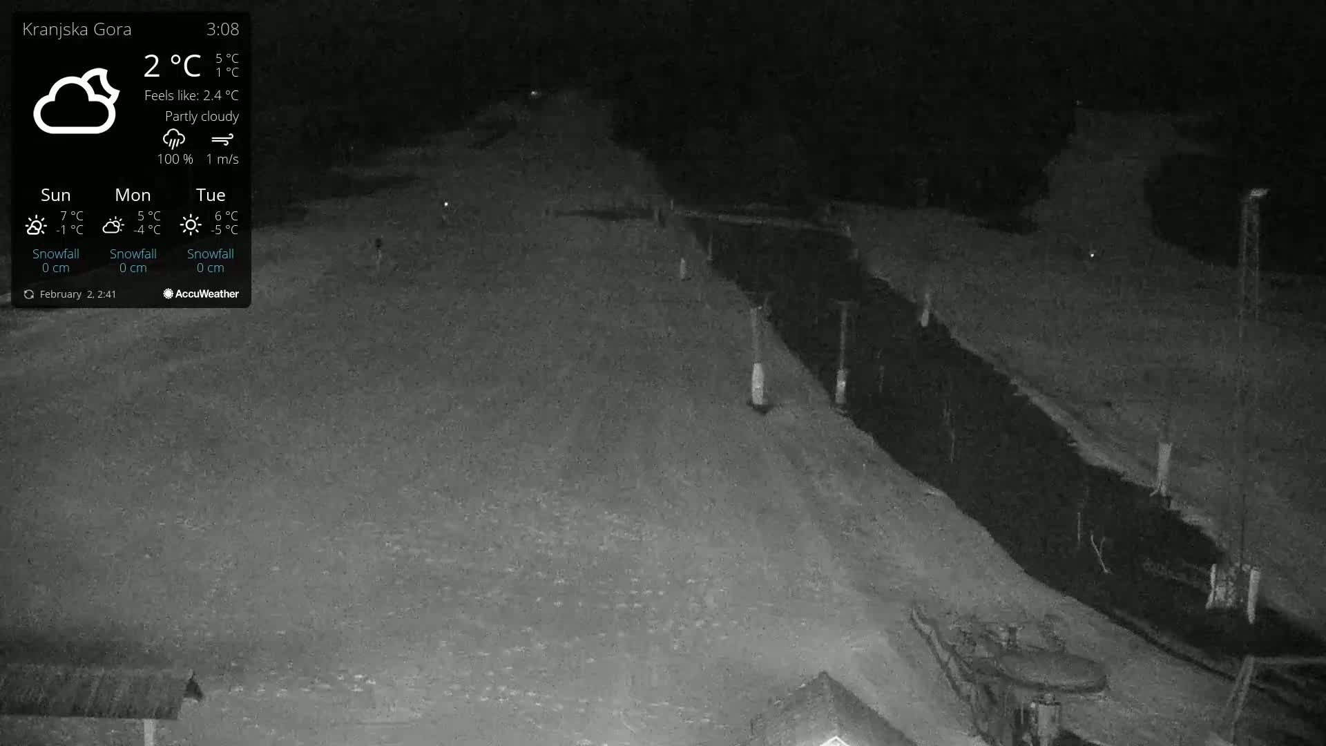 A nighttime, partly cloudy scene shows a snow-covered slope next to a dark, paved area.