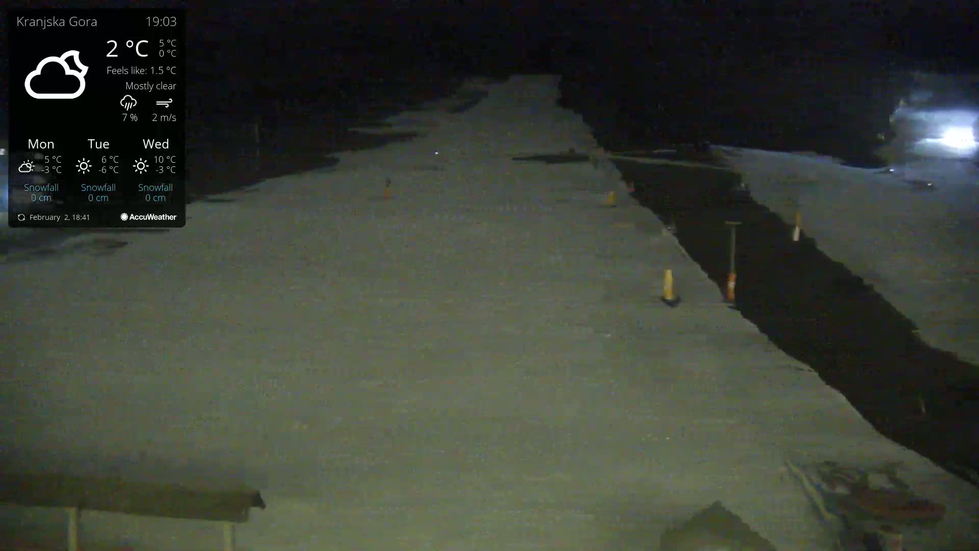 A mostly clear night scene shows a snow-covered area with a dark, possibly paved, section running through it, marked by orange cones and tools.