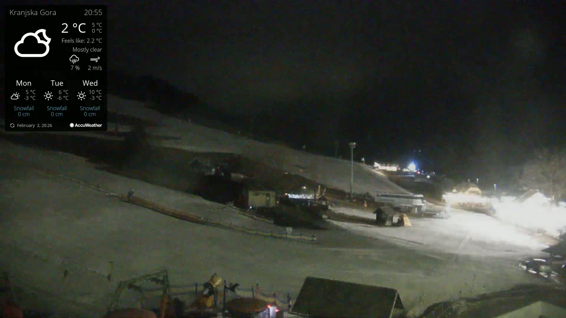 A mostly clear night scene shows a snow-covered ski slope with some lights and equipment visible under the night sky.