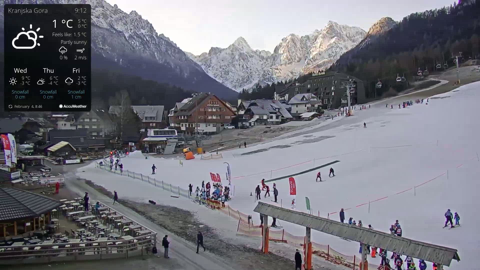 A partly sunny mountain ski slope with several skiers and snowboarders, a village of buildings, and snow-covered mountains in the background.