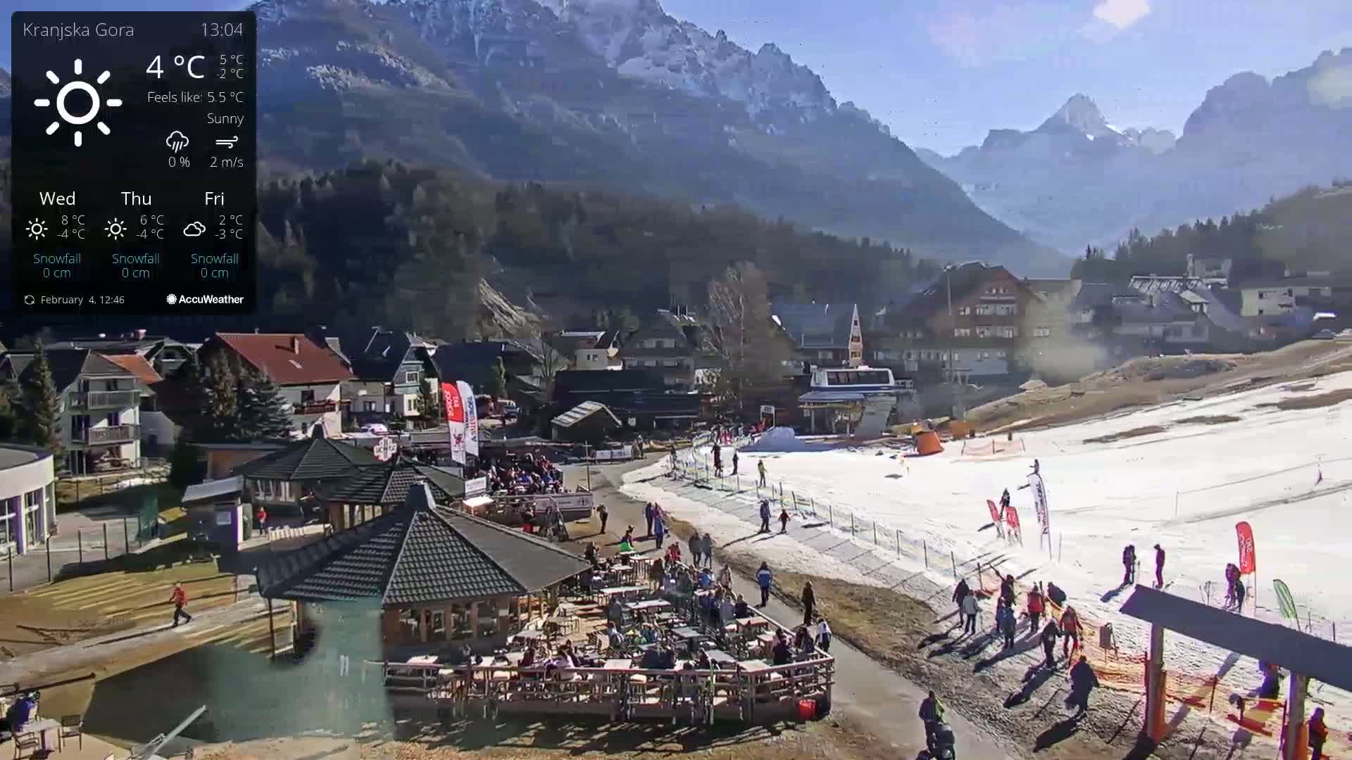 A sunny mountain village scene shows a partially snow-covered ski slope with people walking and sitting at outdoor cafes, all set against a backdrop of snow-capped mountains.