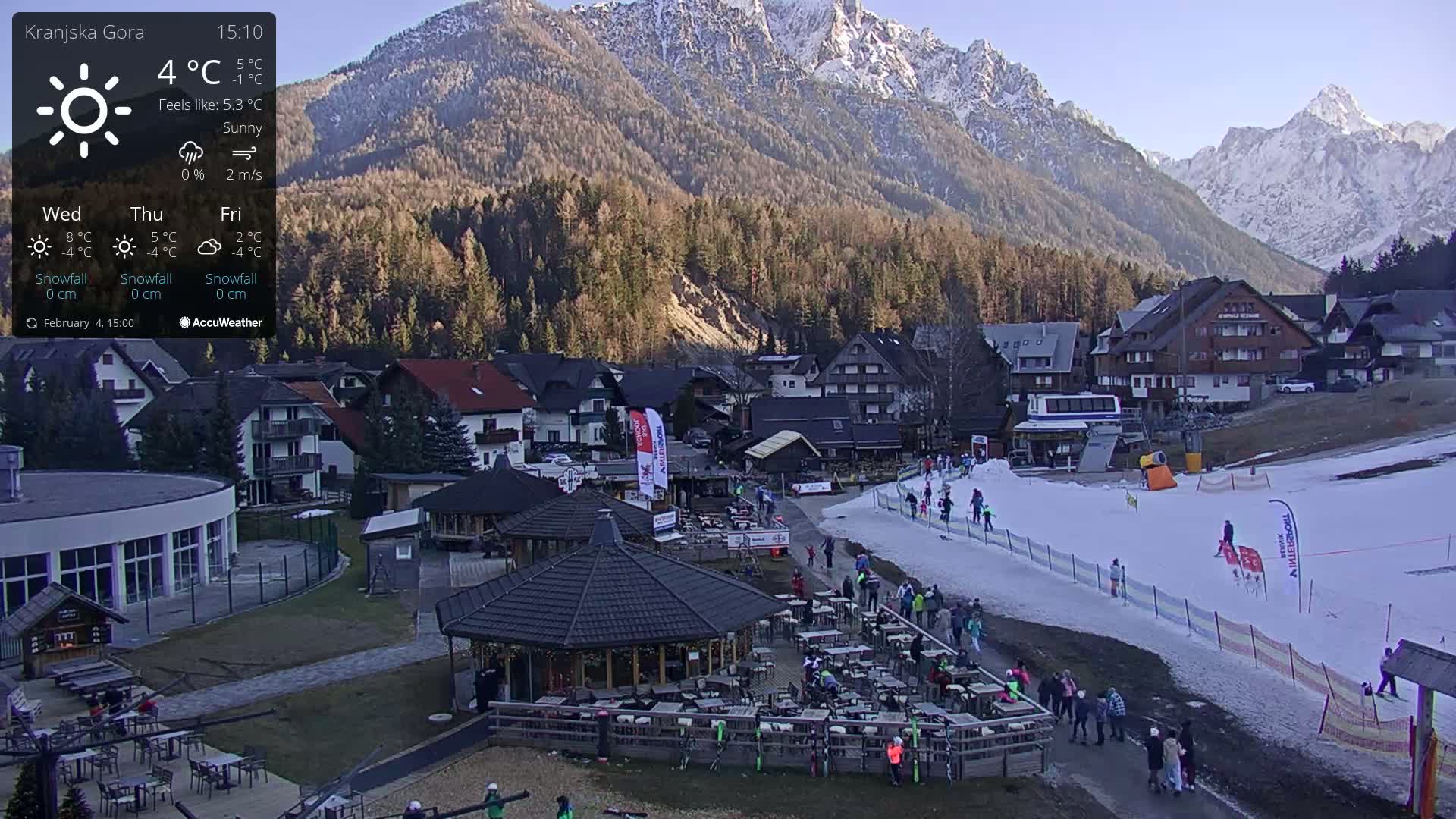 A sunny mountain village with a small ski slope and outdoor seating at a restaurant, nestled in a valley with snow-capped mountains in the background.