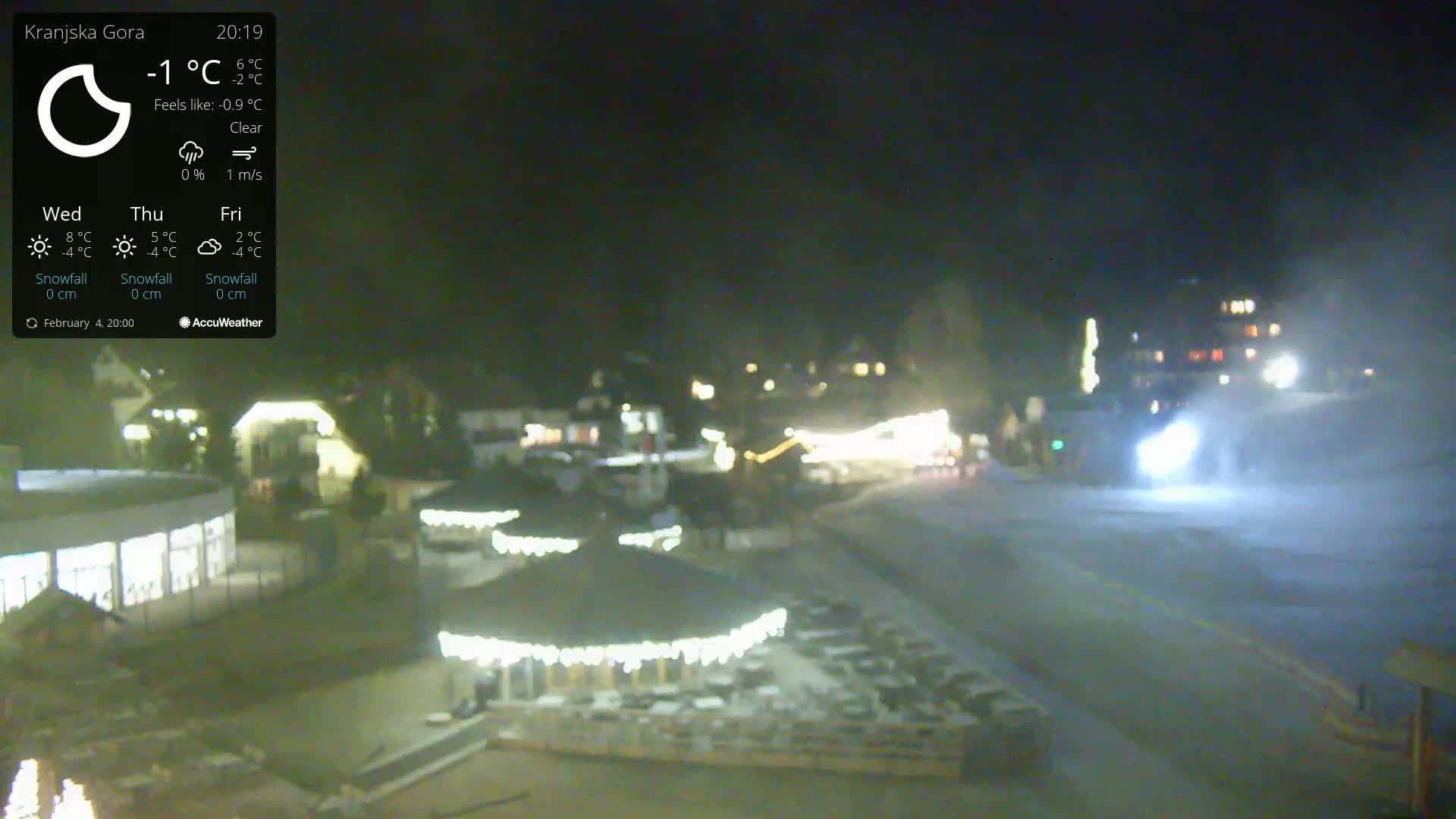 A nighttime, clear, snowy scene shows a snow-covered town square with illuminated buildings and outdoor structures.