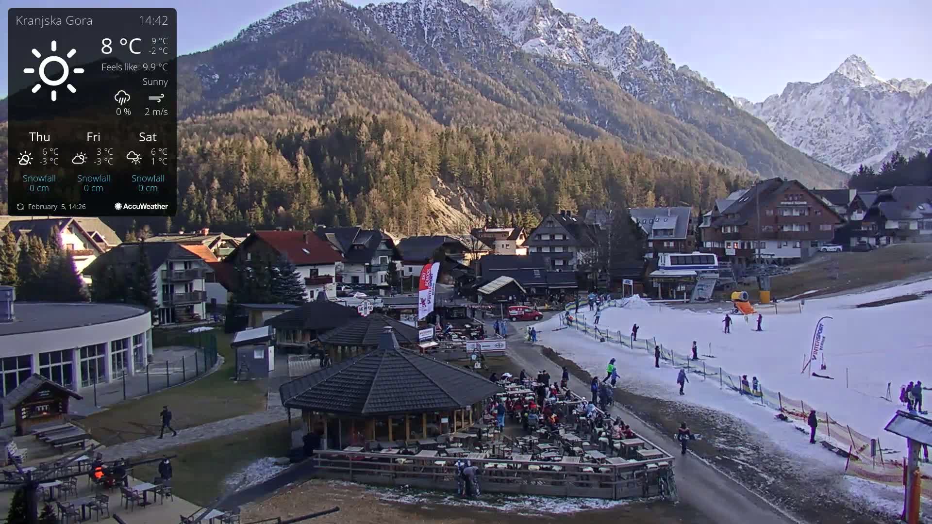 A sunny mountain village with a ski slope and outdoor seating at a restaurant, under a partly cloudy sky.