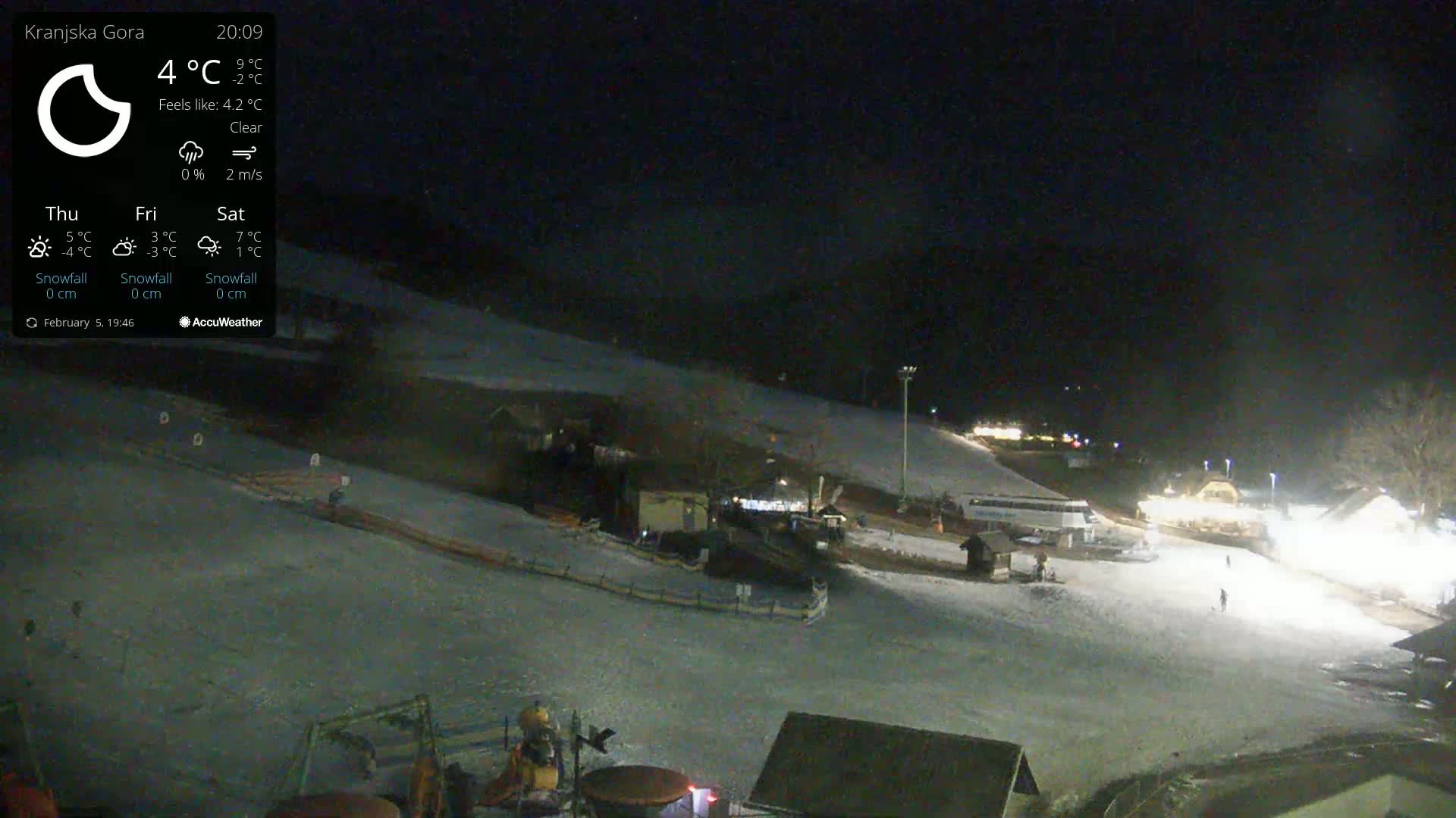 A nighttime, clear view of a snow-covered ski slope with several buildings and a few people visible under artificial lights.