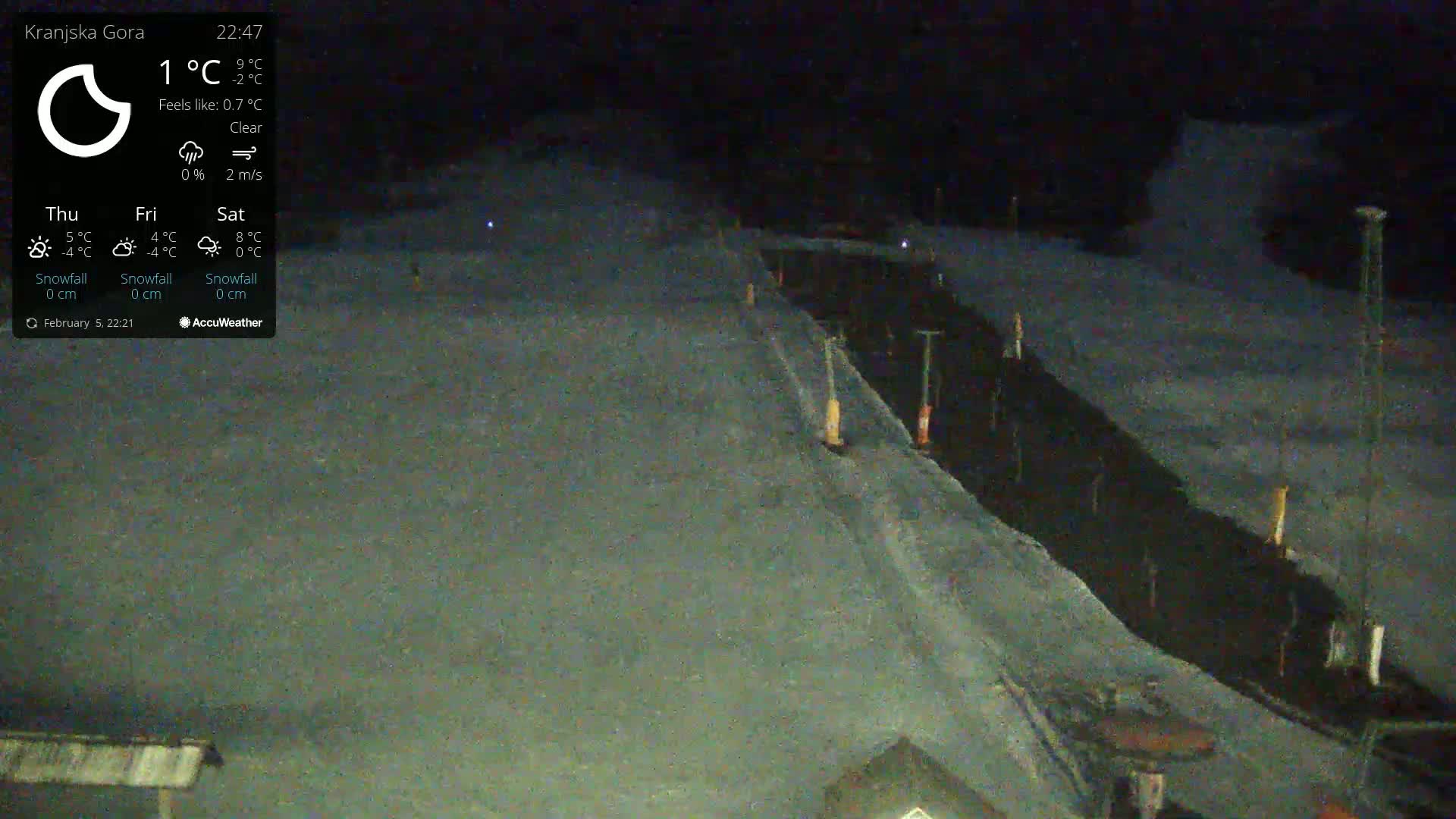 A nighttime, clear-weather view of a snow-covered slope with a dark path or road running down the center, marked by short posts.