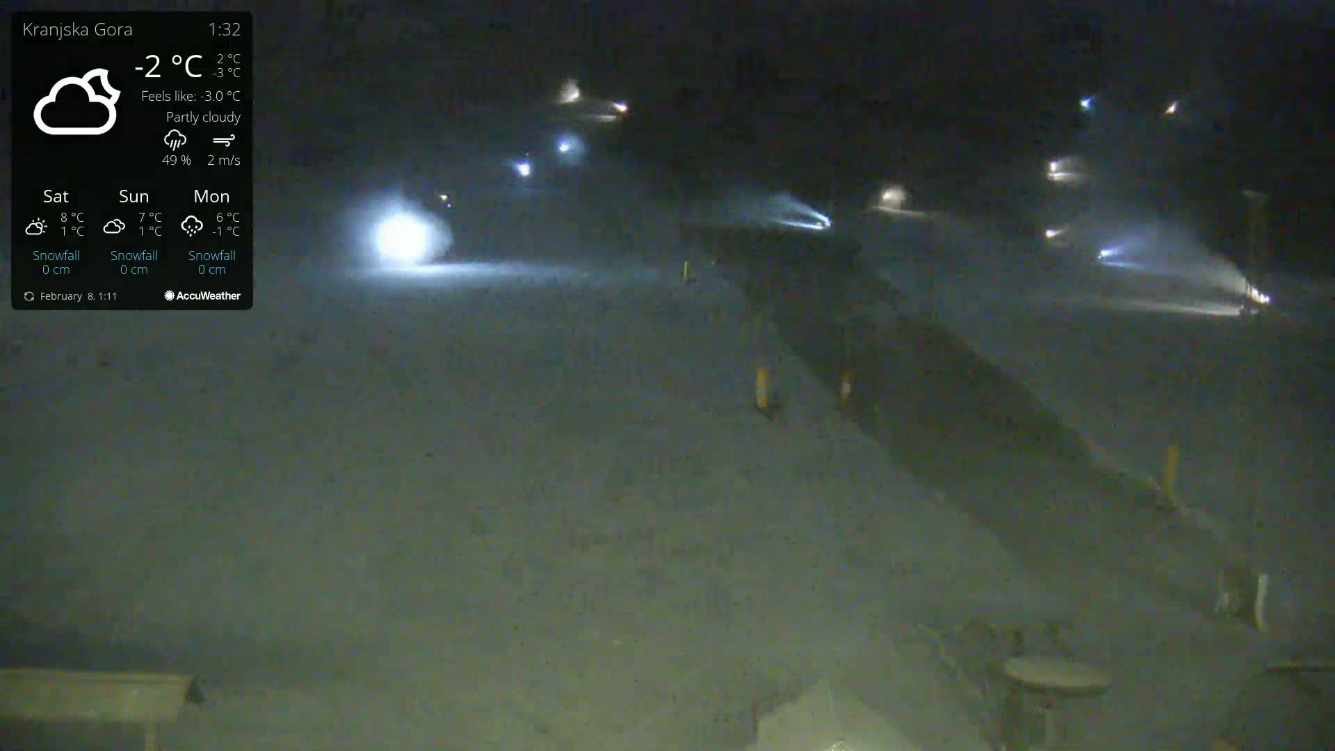 A nighttime, partly cloudy scene shows a snow-covered area with several vehicles’ headlights visible in the distance.