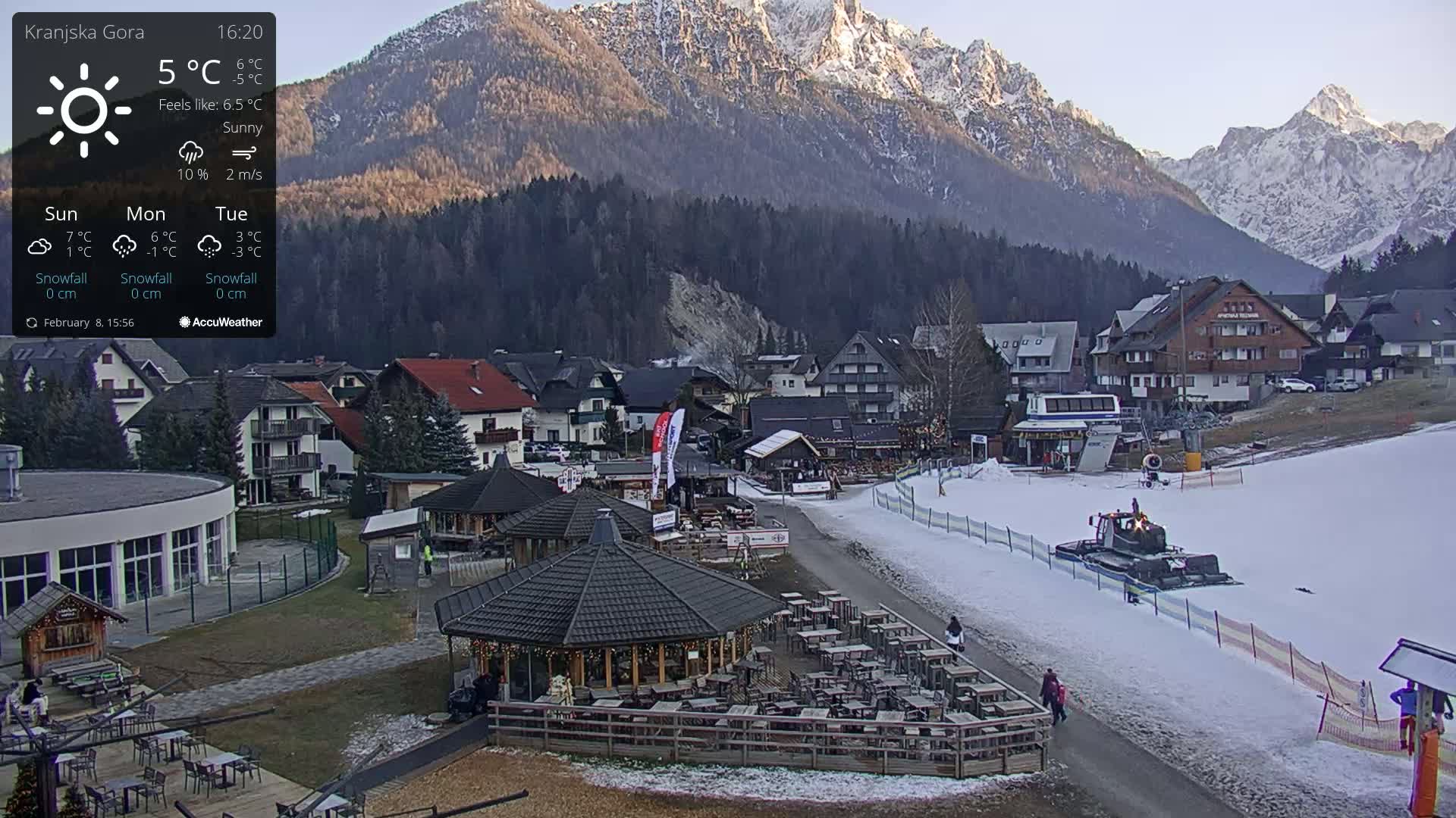 A sunny mountain village with snow on the ground and a snow groomer on a ski slope, viewed from above.