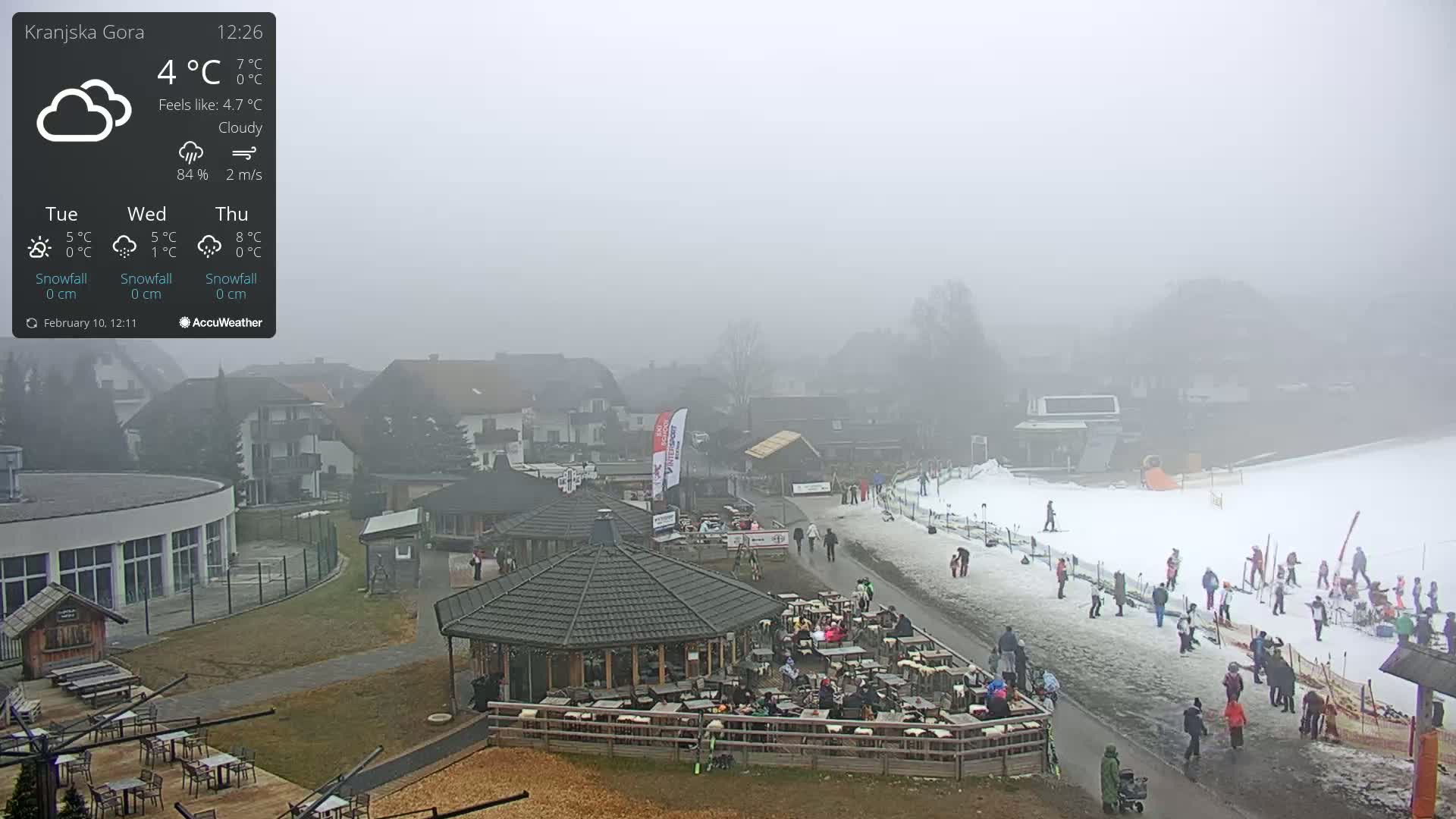 A foggy, cloudy outdoor scene shows a ski resort with people gathered around outdoor seating areas near a small ski slope.