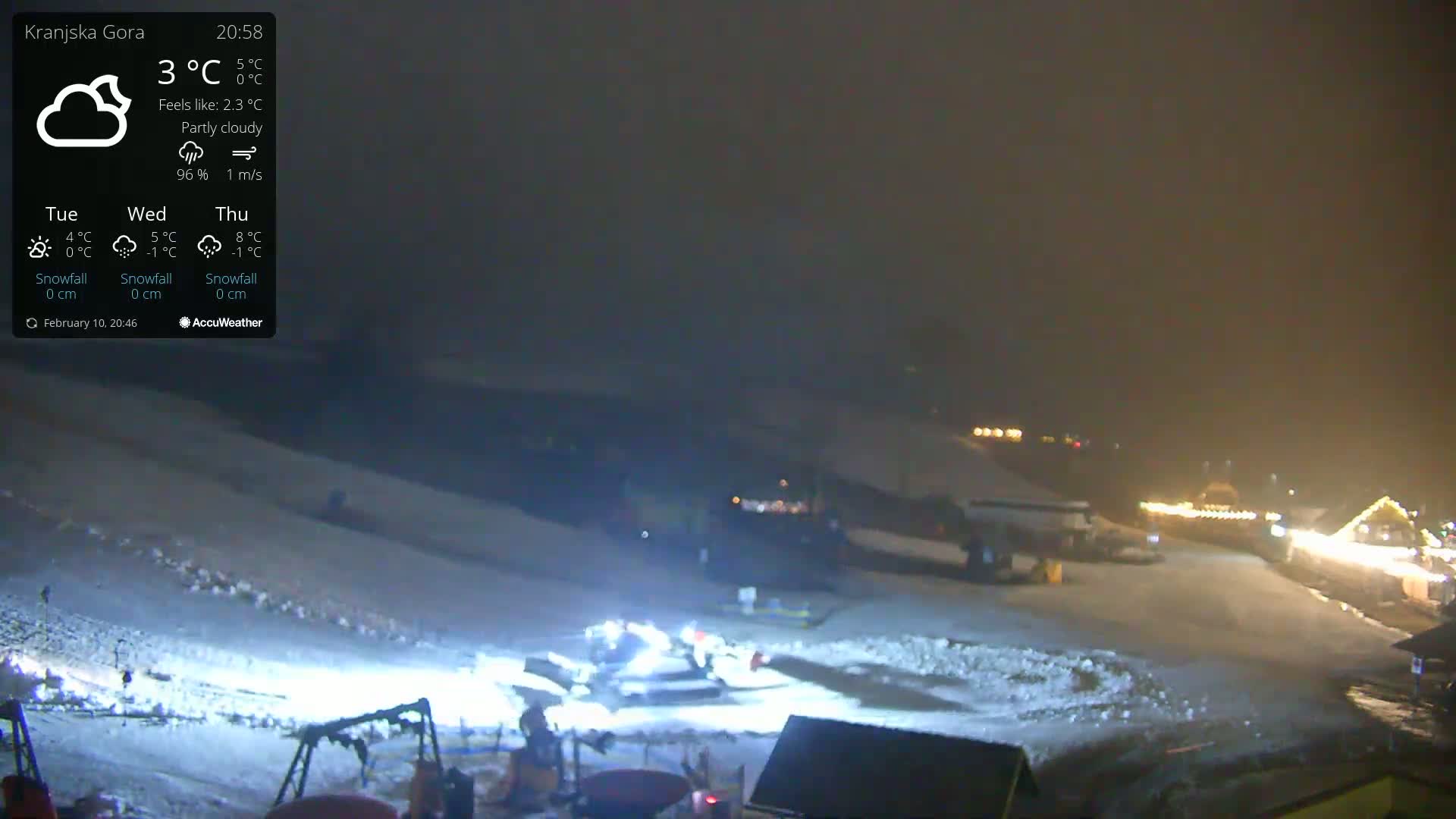 A snow-covered slope at night under partly cloudy skies, illuminated by lights from buildings in the distance and bright lights from snow-grooming equipment.