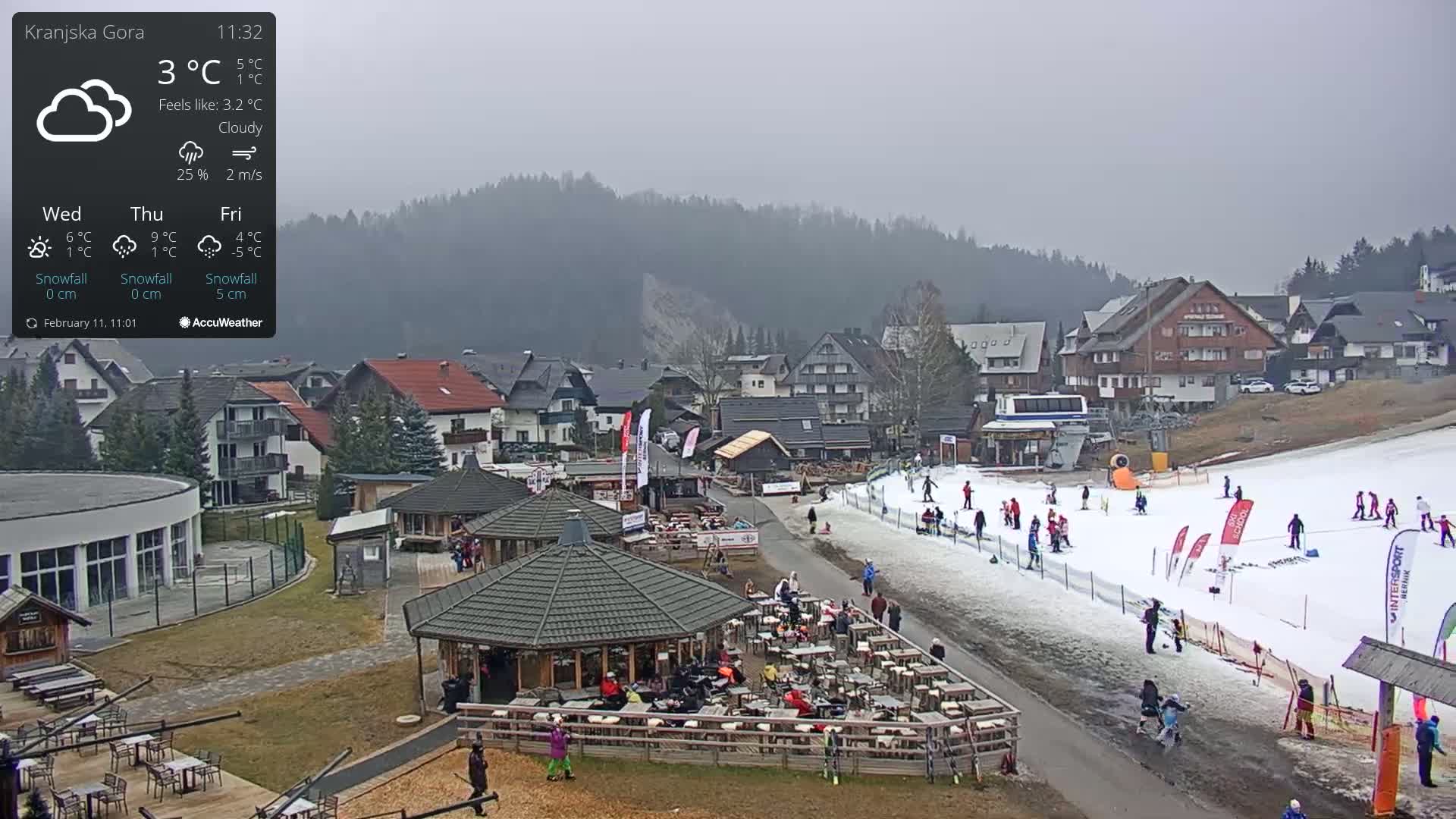 A cloudy day in a ski resort town shows a small ski slope with several people skiing and a patio area with many people seated at tables.