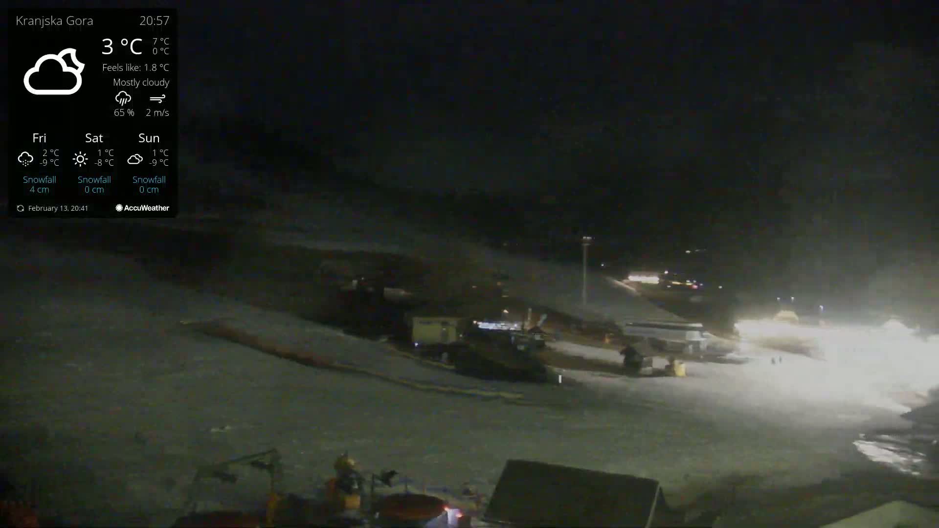 A mostly cloudy night scene shows a snow-covered slope with several vehicles and lights visible in the distance.