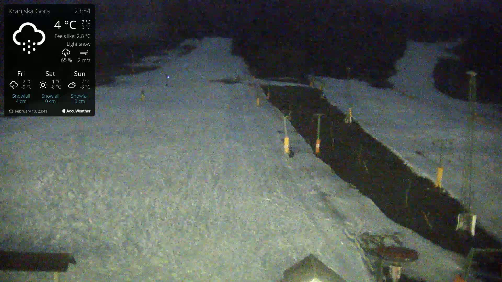 A nighttime, snow-covered ski slope with a dark, narrow path running through it under light snow.