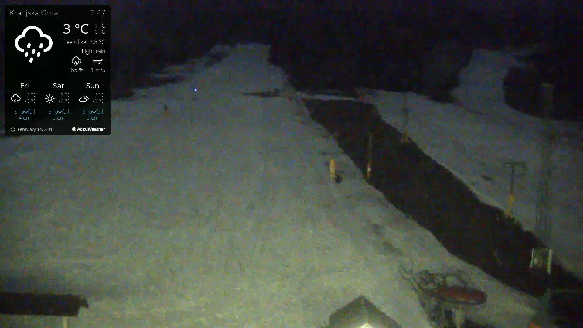 A snow-covered area with a dark, possibly paved, path running through it under light rain at night.