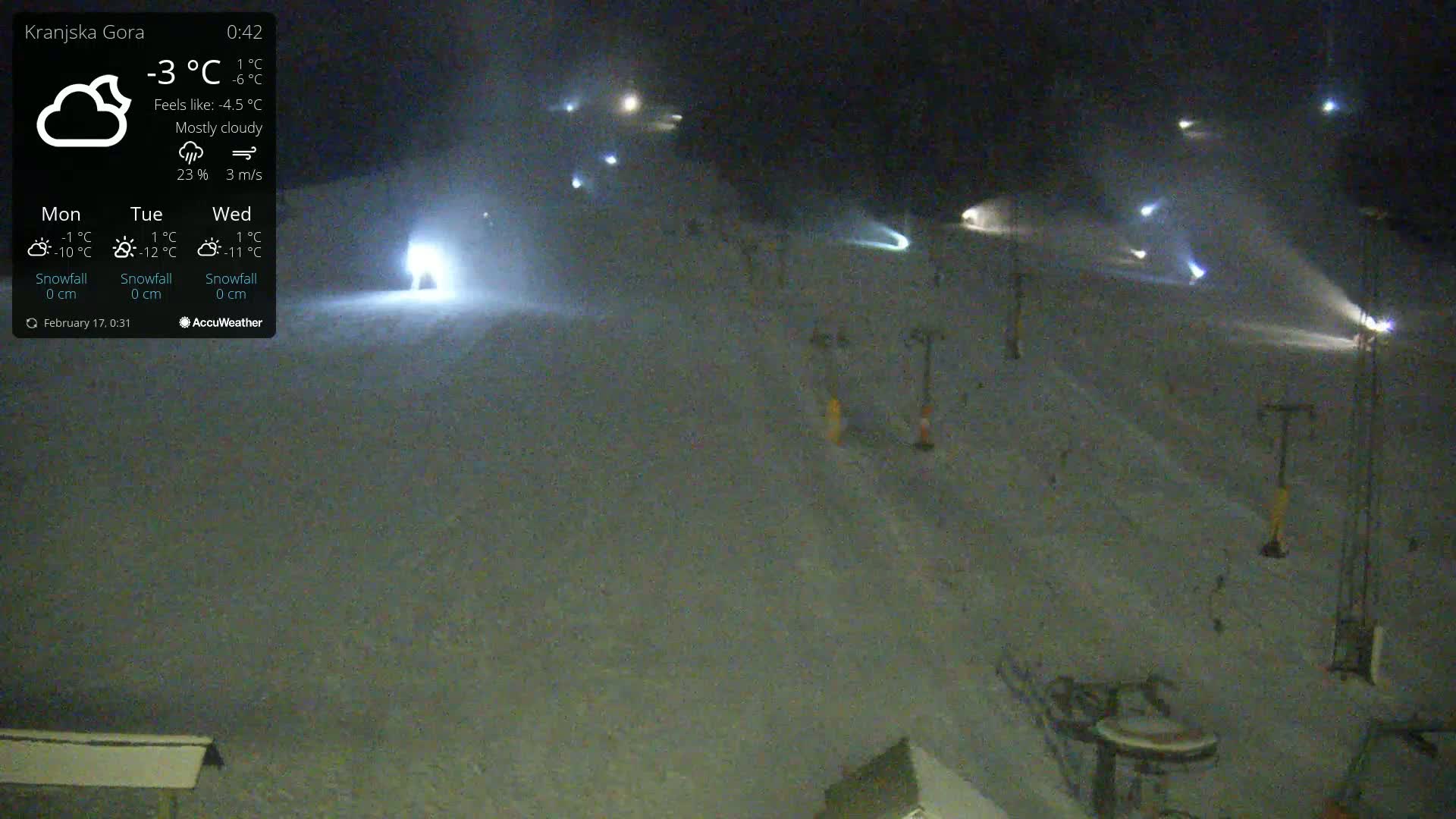 A nighttime, mostly cloudy view of a snow-covered ski slope with several lights illuminating the area and a few ski lifts visible.