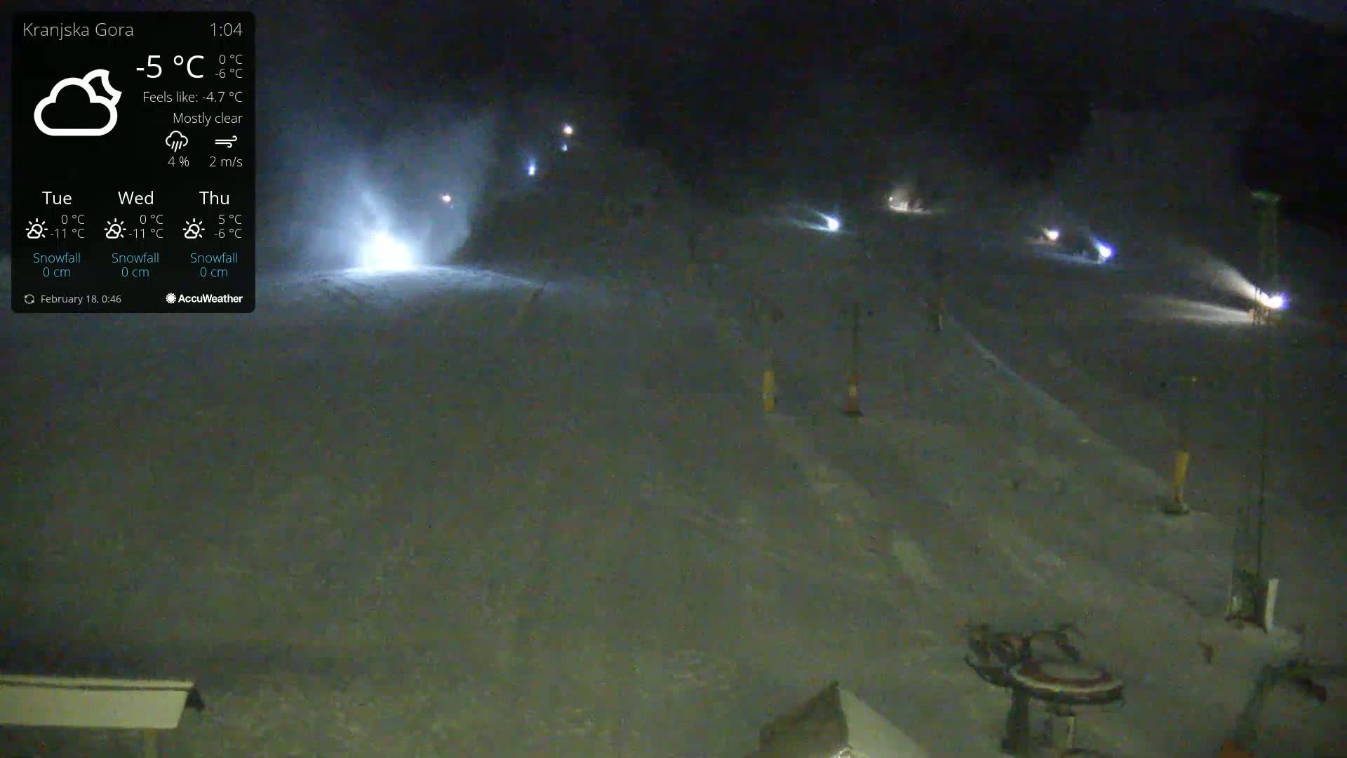 A nighttime, mostly clear view of a snow-covered slope with several lights and some activity visible under the lights; a light snow is falling.