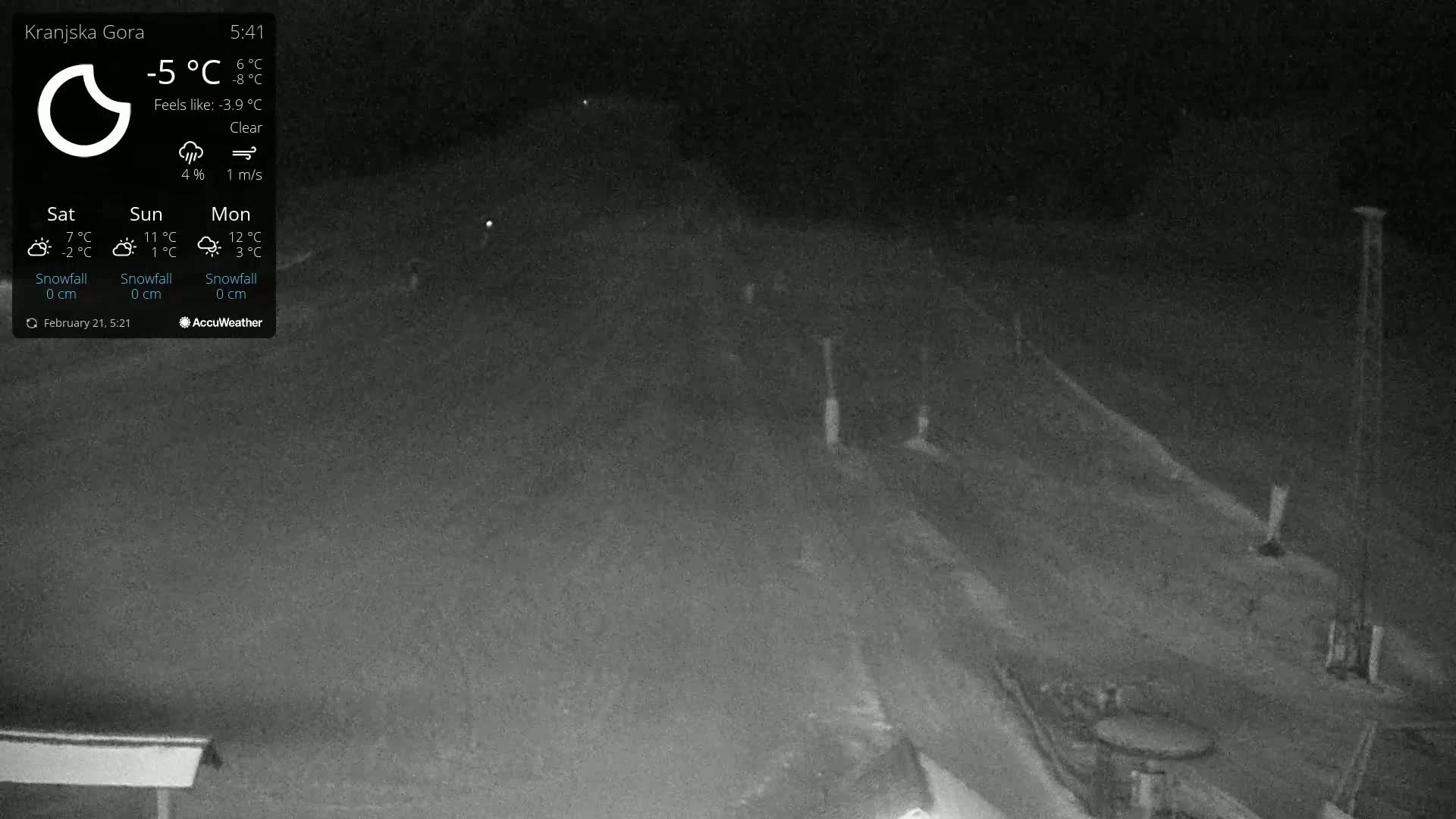 A nighttime, clear-weather view of a snow-covered road with some indistinct structures and objects visible in the distance.