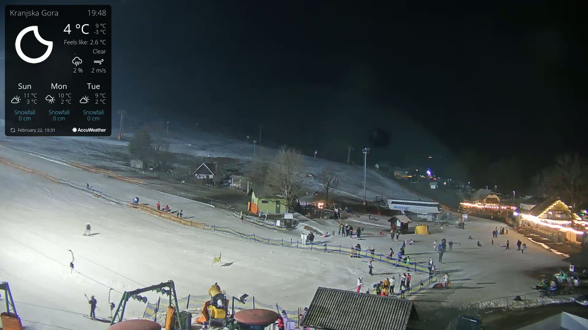 A nighttime view of a snow-covered ski slope with people skiing and milling around near illuminated buildings under a clear sky.