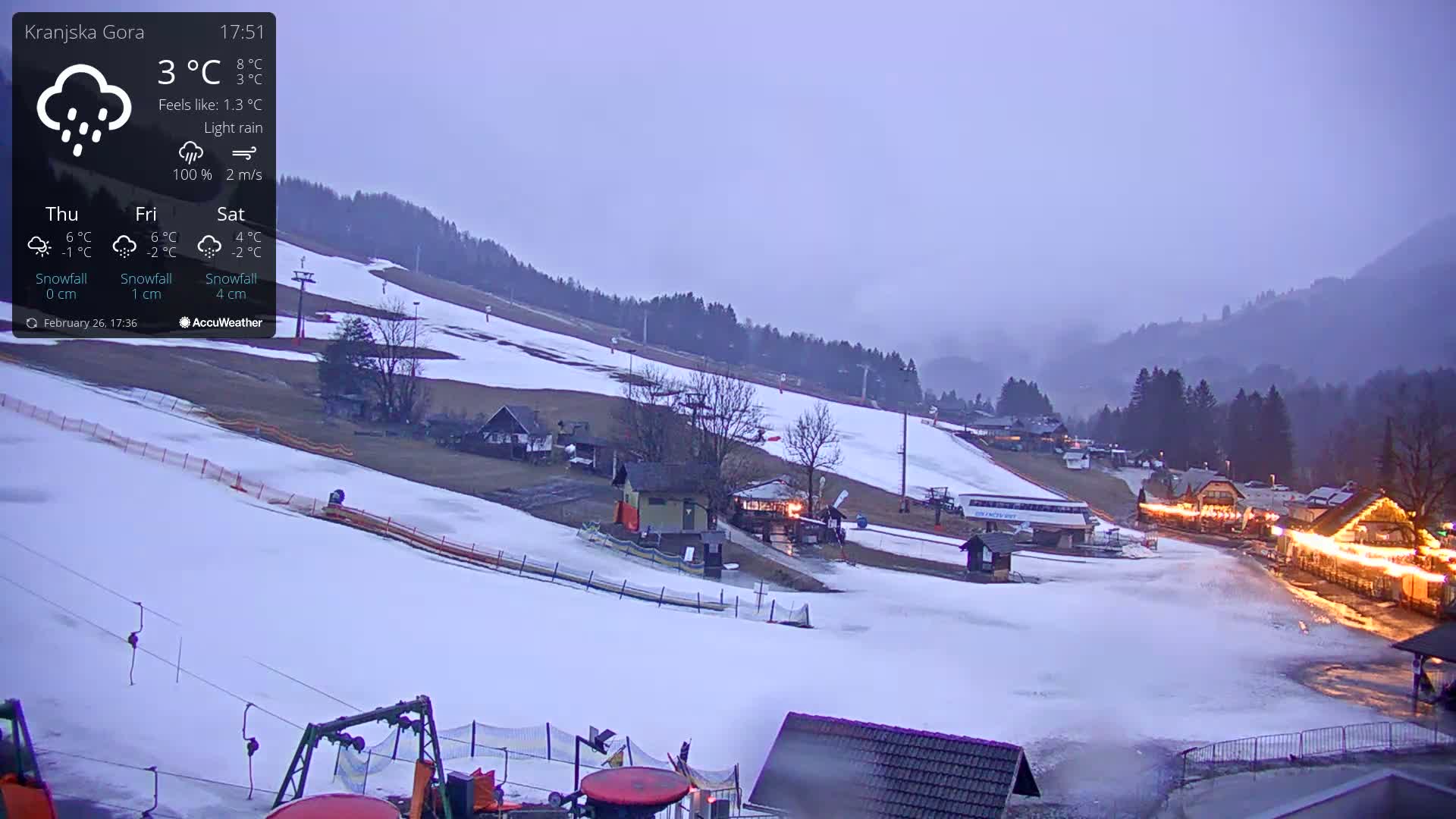 A snow-covered ski slope with buildings and lights at the base under a light rain and overcast sky.