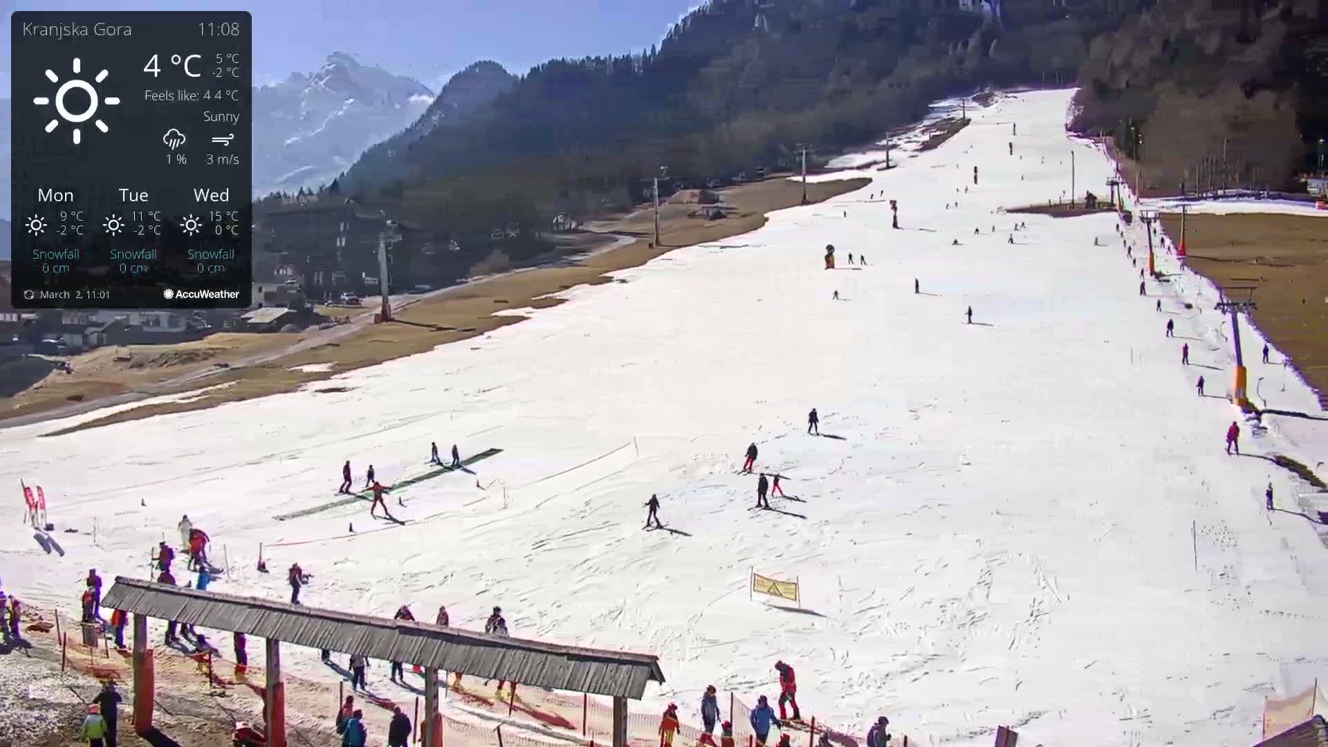 A sunny ski slope with numerous skiers and snowboarders on a snow-covered hill, backed by mountains.