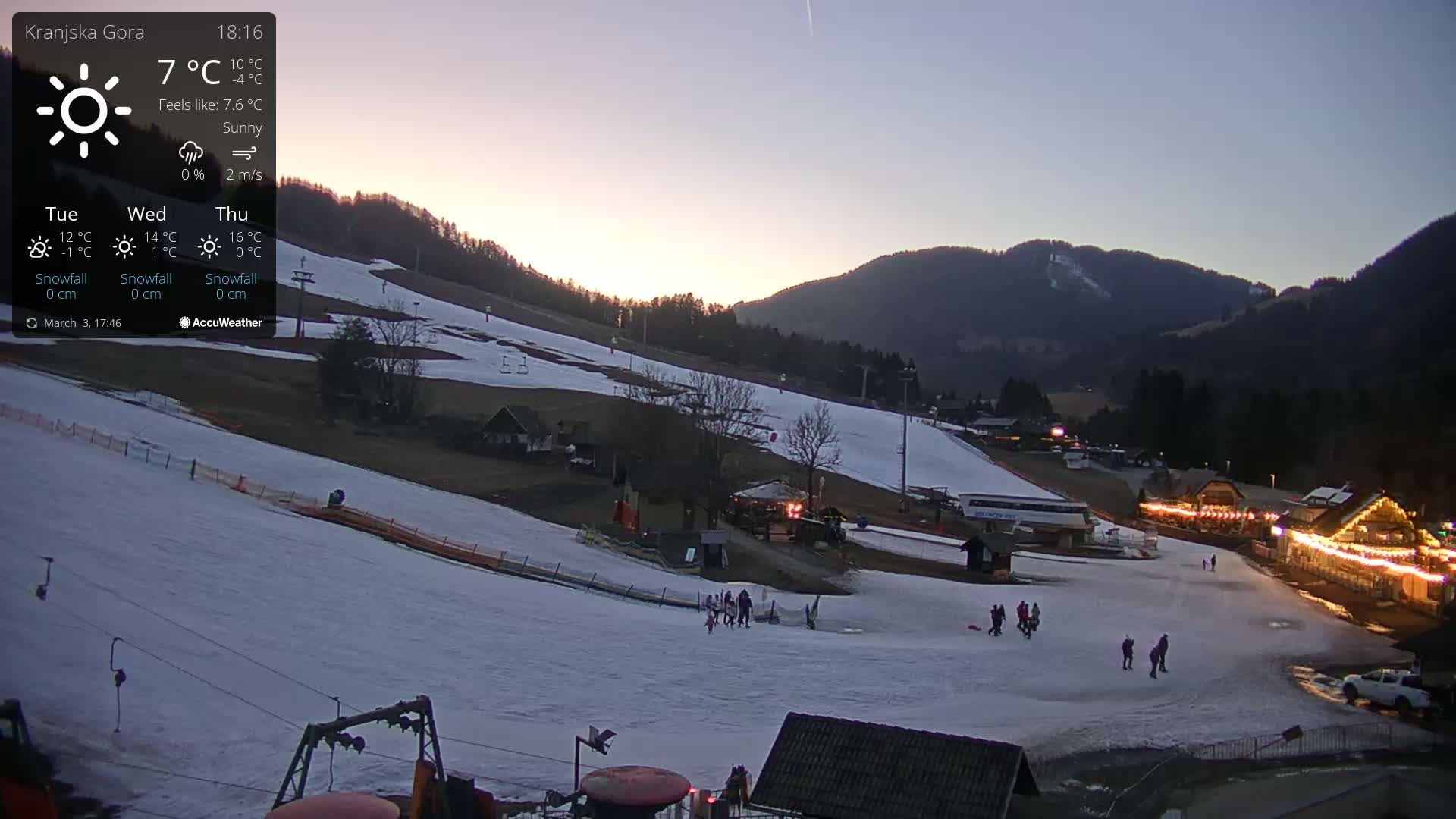 A snowy ski slope at dusk, with several people walking near the base of the hill and buildings illuminated with lights.