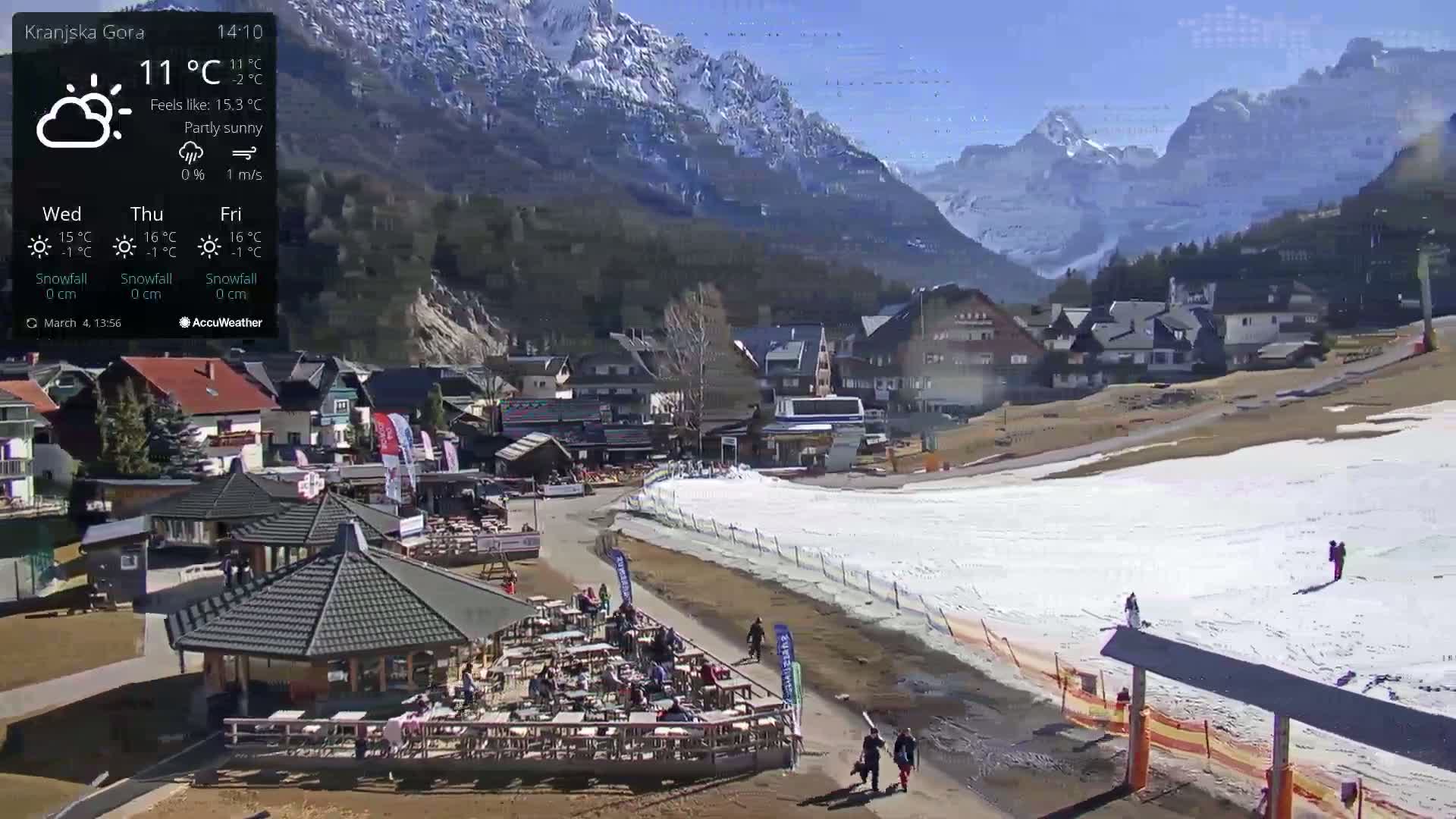 A partly sunny mountain village with a snow-covered ski slope shows several people walking near outdoor seating areas, with snow-capped mountains in the background.