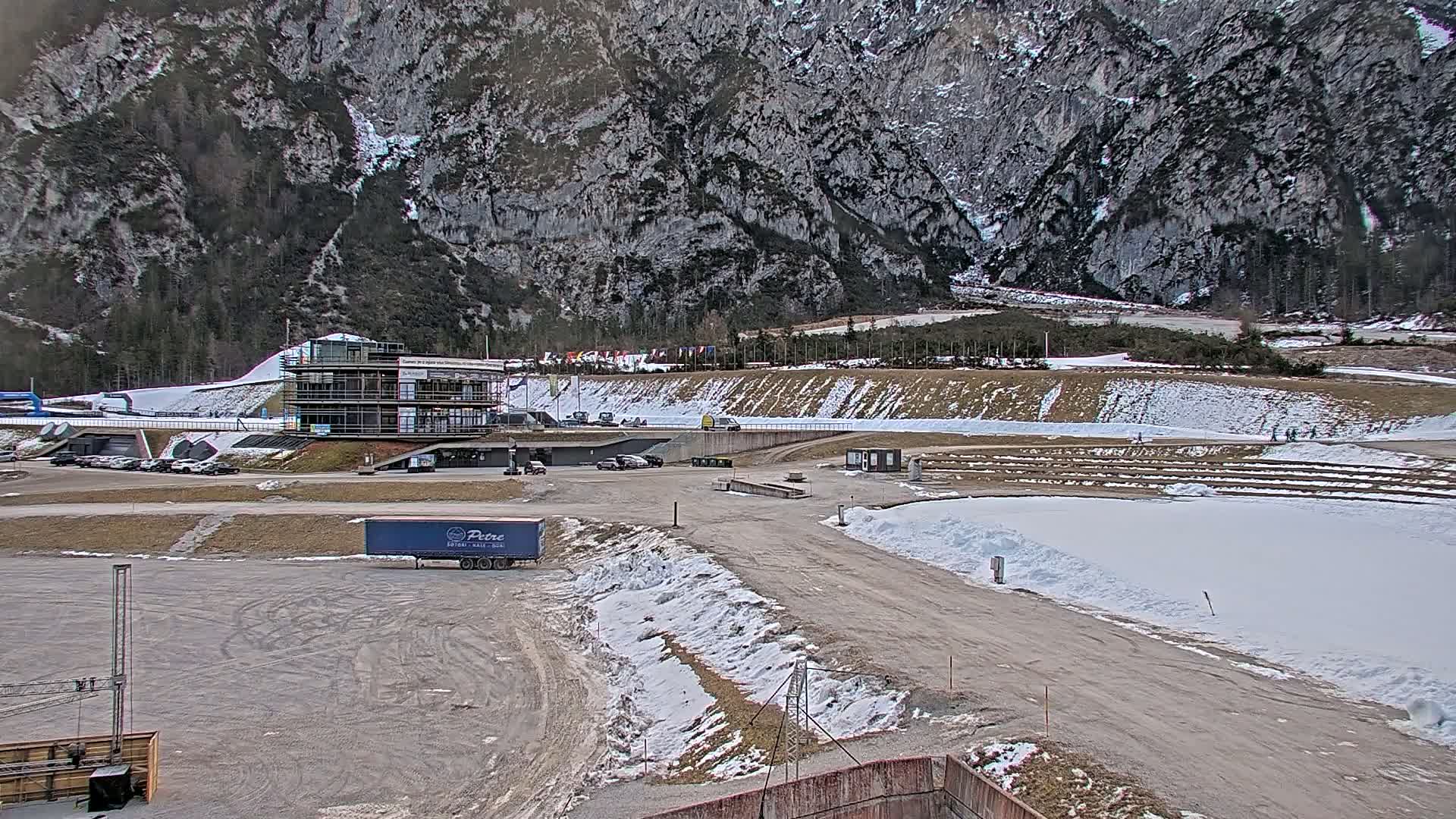 A snow-covered outdoor area with a building, vehicles, and a large rocky mountain in the background.