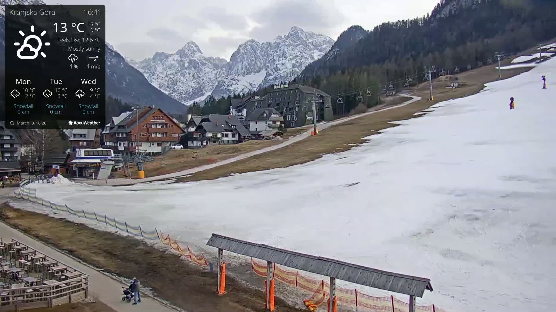 A mostly sunny mountain village scene with patchy snow on the ground, a ski lift, and snow-capped mountains in the background.