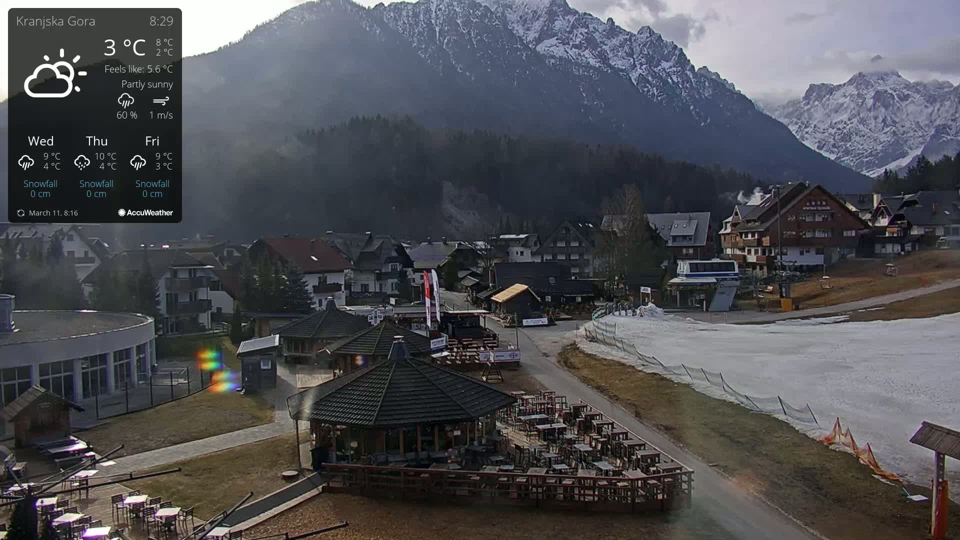 A partly sunny mountain village with a ski area, featuring outdoor seating at a restaurant, under a mostly clear sky.