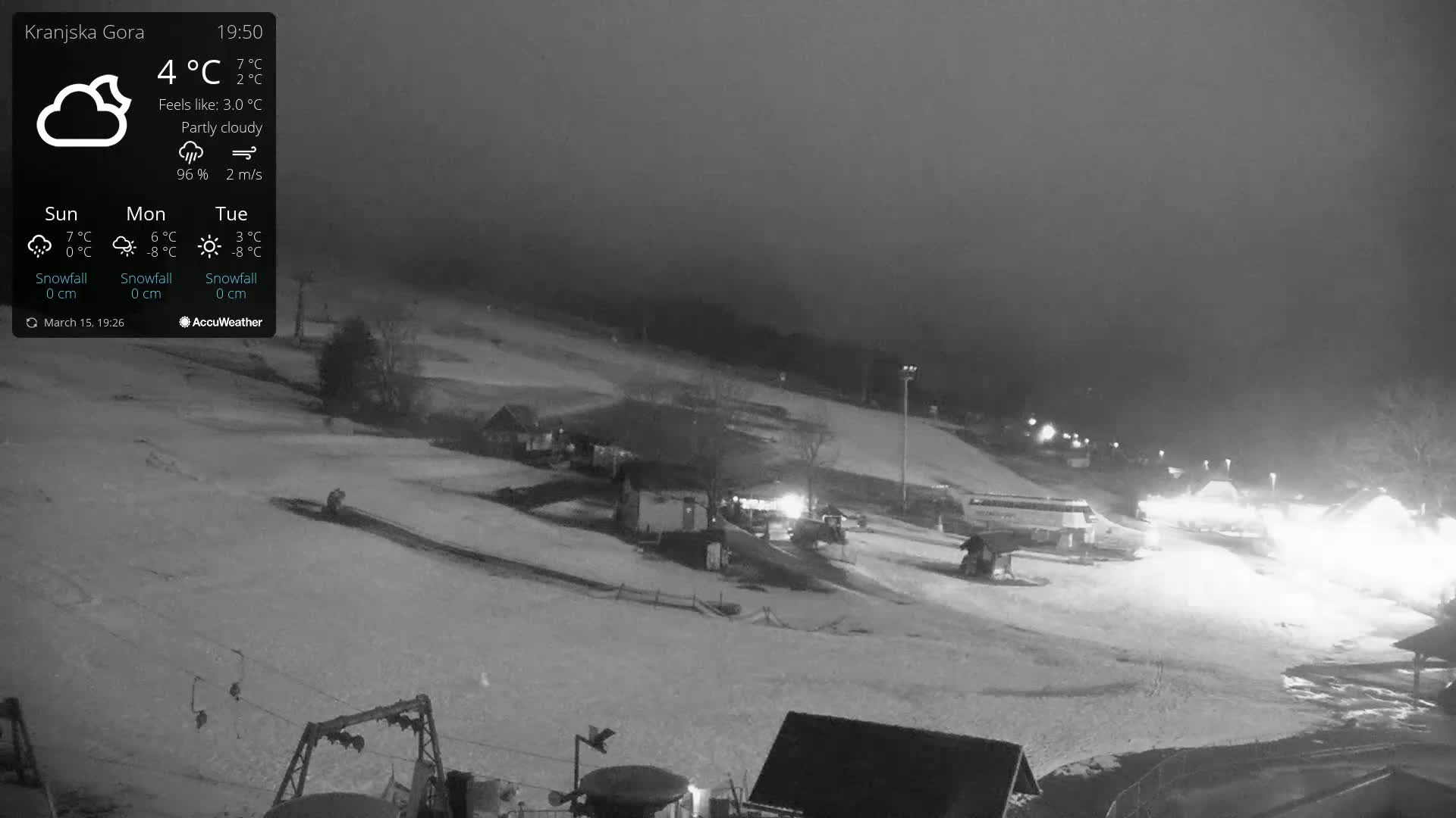 A nighttime, partly cloudy view of a snow-covered ski slope with some buildings and vehicles visible under dim lighting.