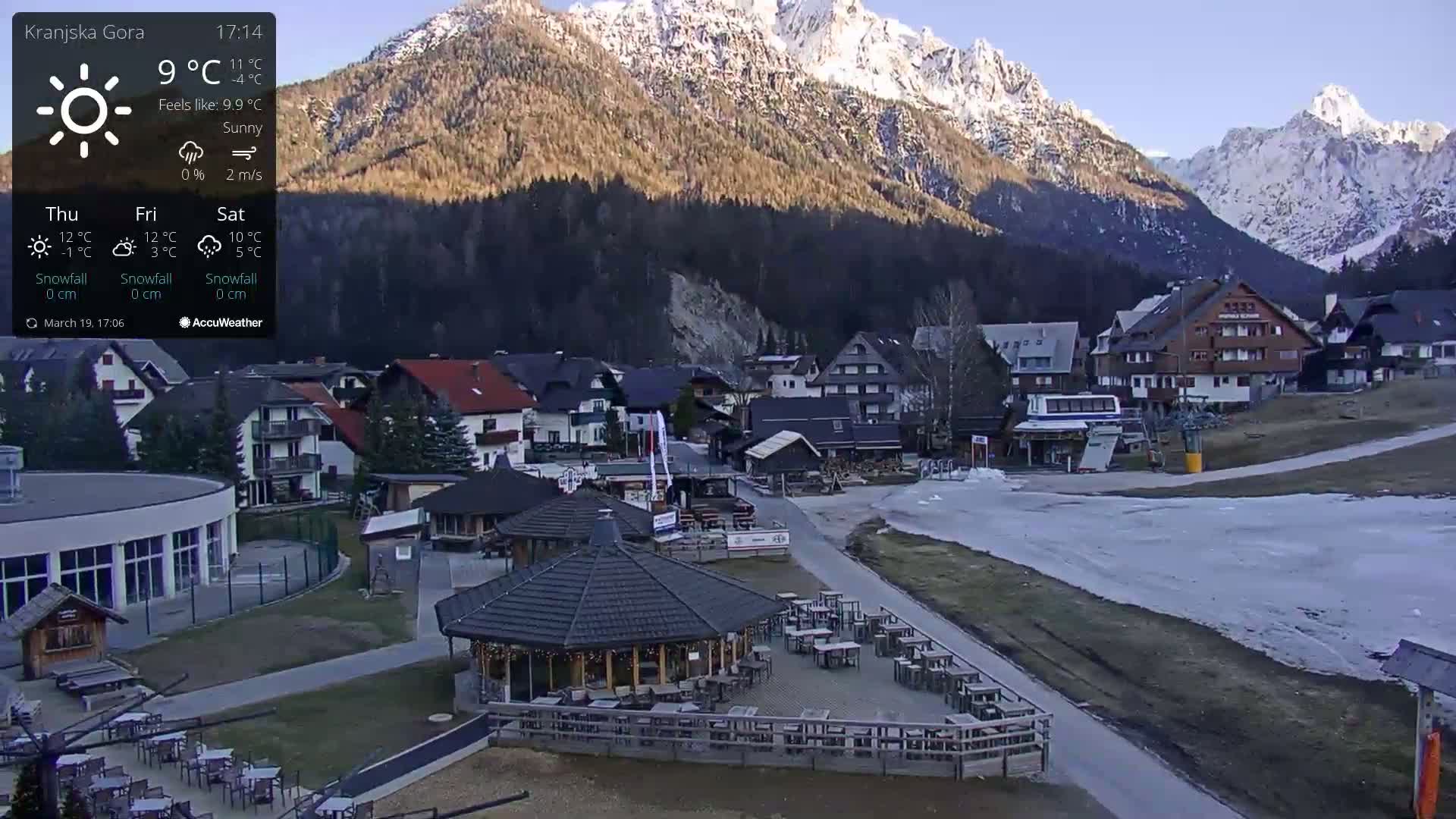 A sunny mountain village with a snow-covered path and a gazebo with outdoor seating, situated at the base of snow-capped mountains.