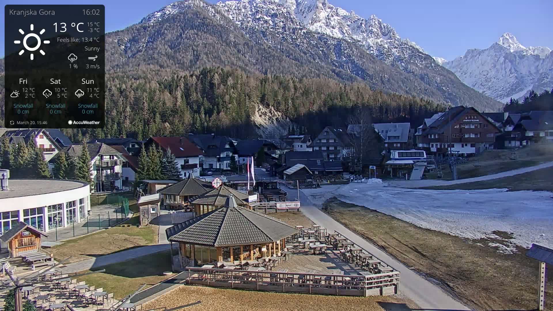 A sunny mountain village with a few buildings, outdoor seating areas, and a ski lift, nestled against a backdrop of snow-capped mountains.