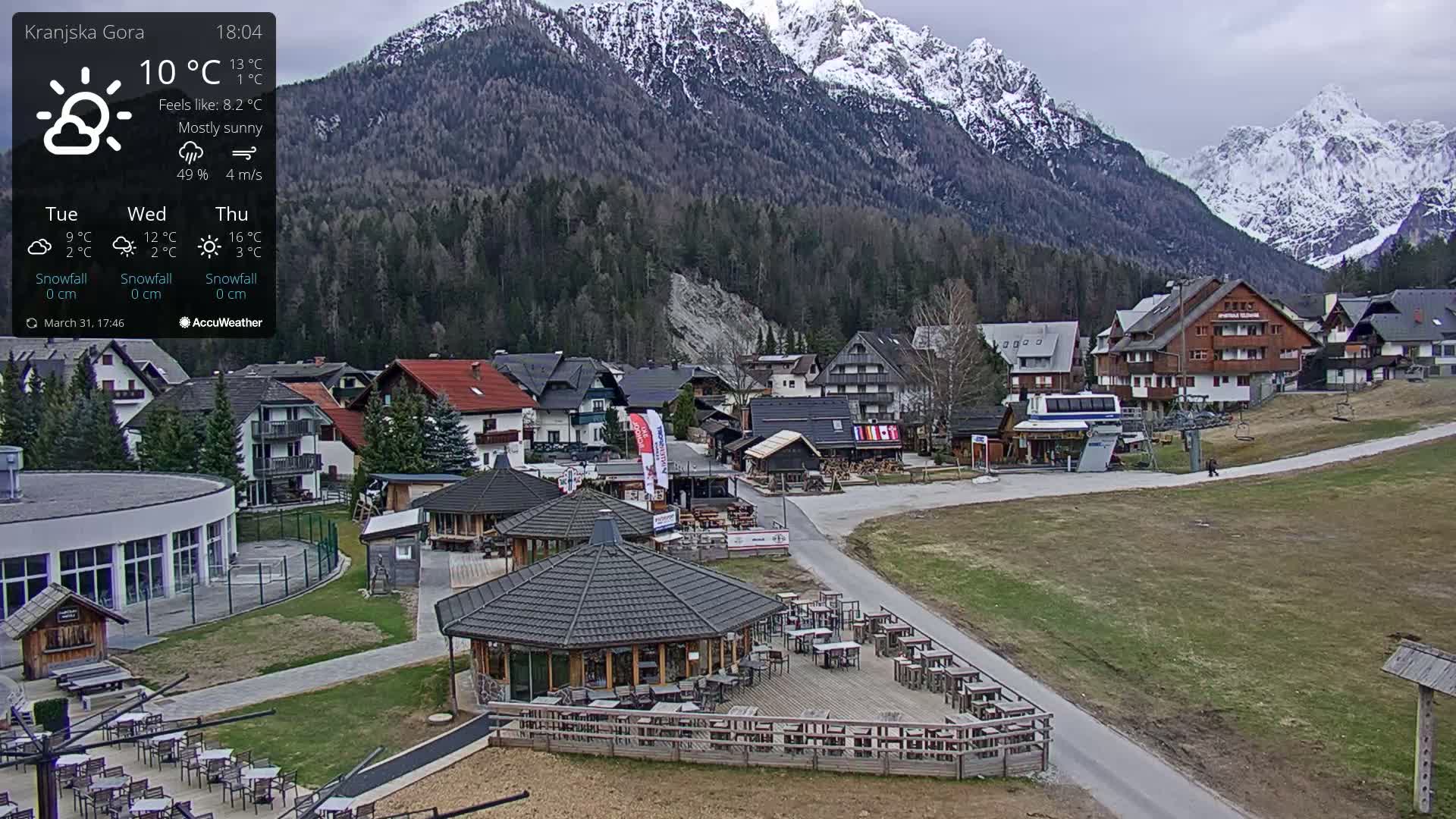 A mostly sunny mountain village with a few buildings, outdoor seating areas, and a chairlift, situated against a backdrop of snow-capped mountains.