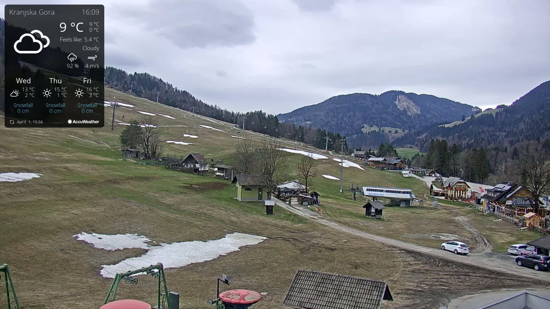A mostly cloudy view of a ski resort with patchy snow on the slopes, some buildings, and a few cars parked in a parking lot.