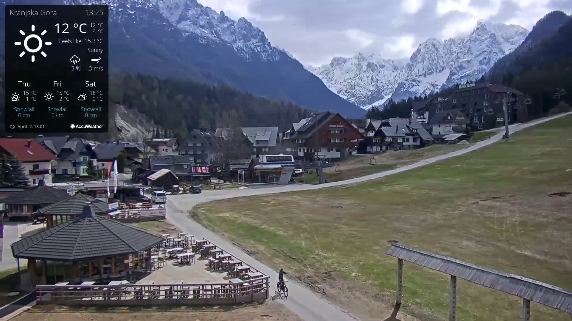 A sunny day reveals a village nestled in a valley, with snow-capped mountains in the background and a cyclist riding on a path near outdoor seating.