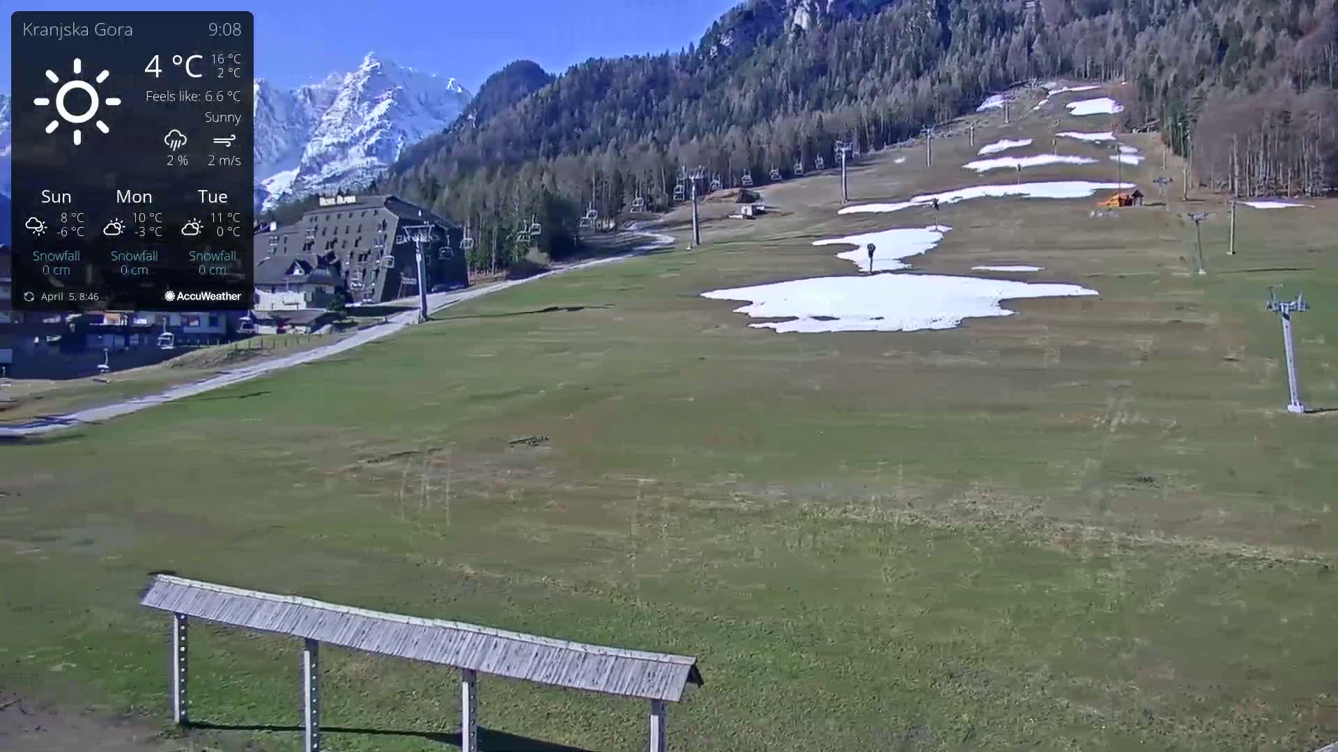 A sunny mountain vista shows a ski resort with patches of remaining snow, chairlifts, and a building, all under a clear blue sky.