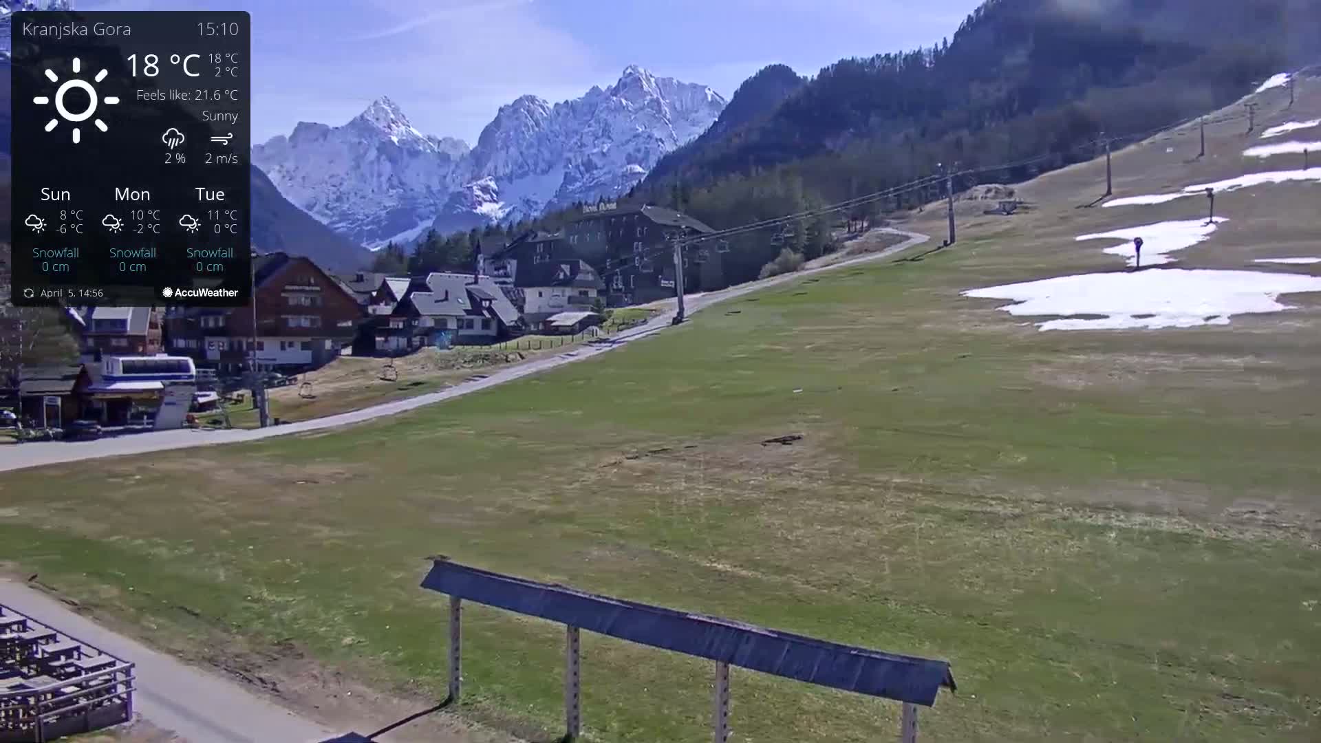 A sunny mountain village scene shows a grassy area with patches of snow, a few buildings, a ski lift, and snow-capped mountains in the background.
