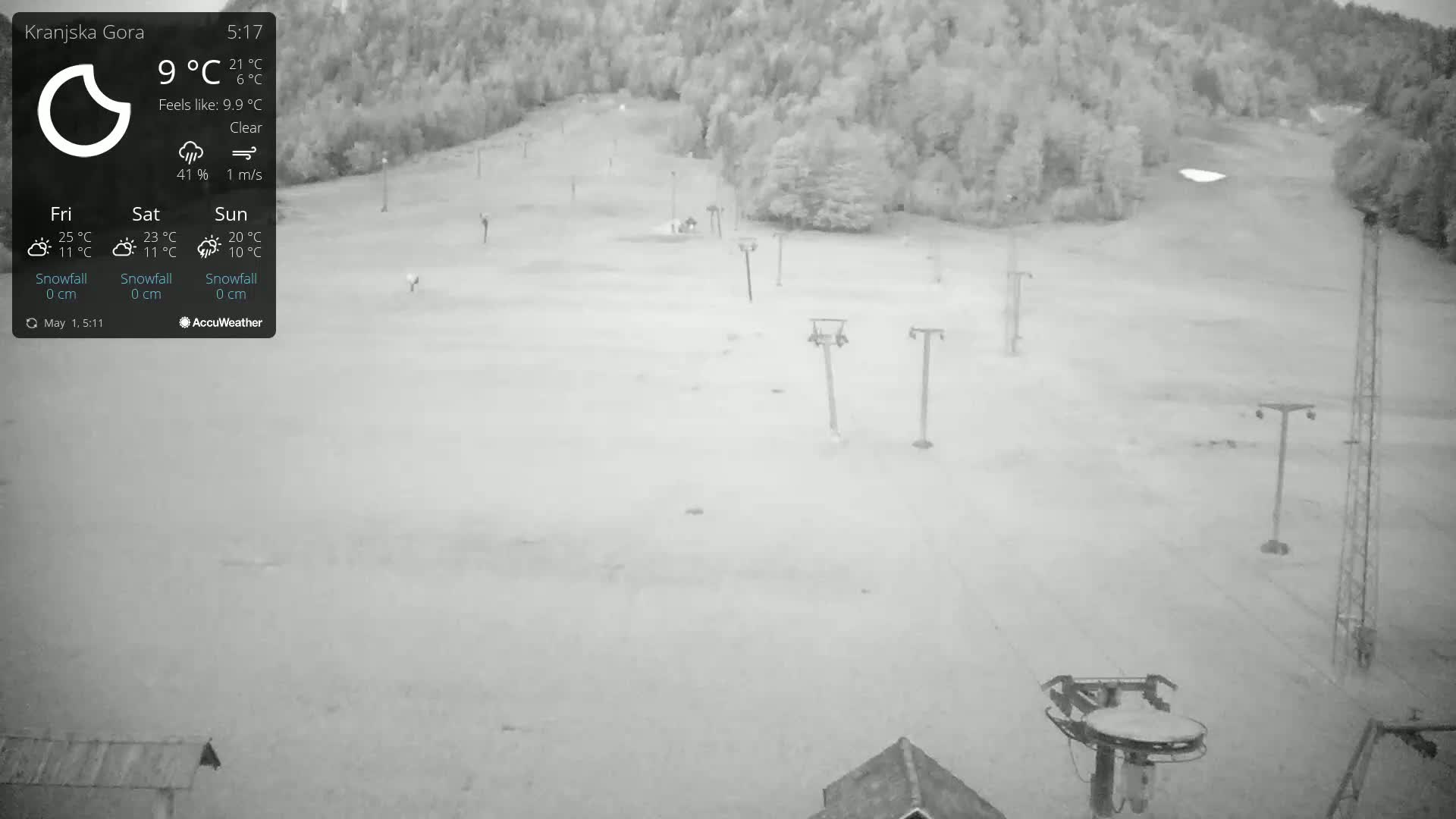 A mostly clear, daytime, high-angle, grayscale view of a ski slope with several chairlift towers and a wooded area in the background.