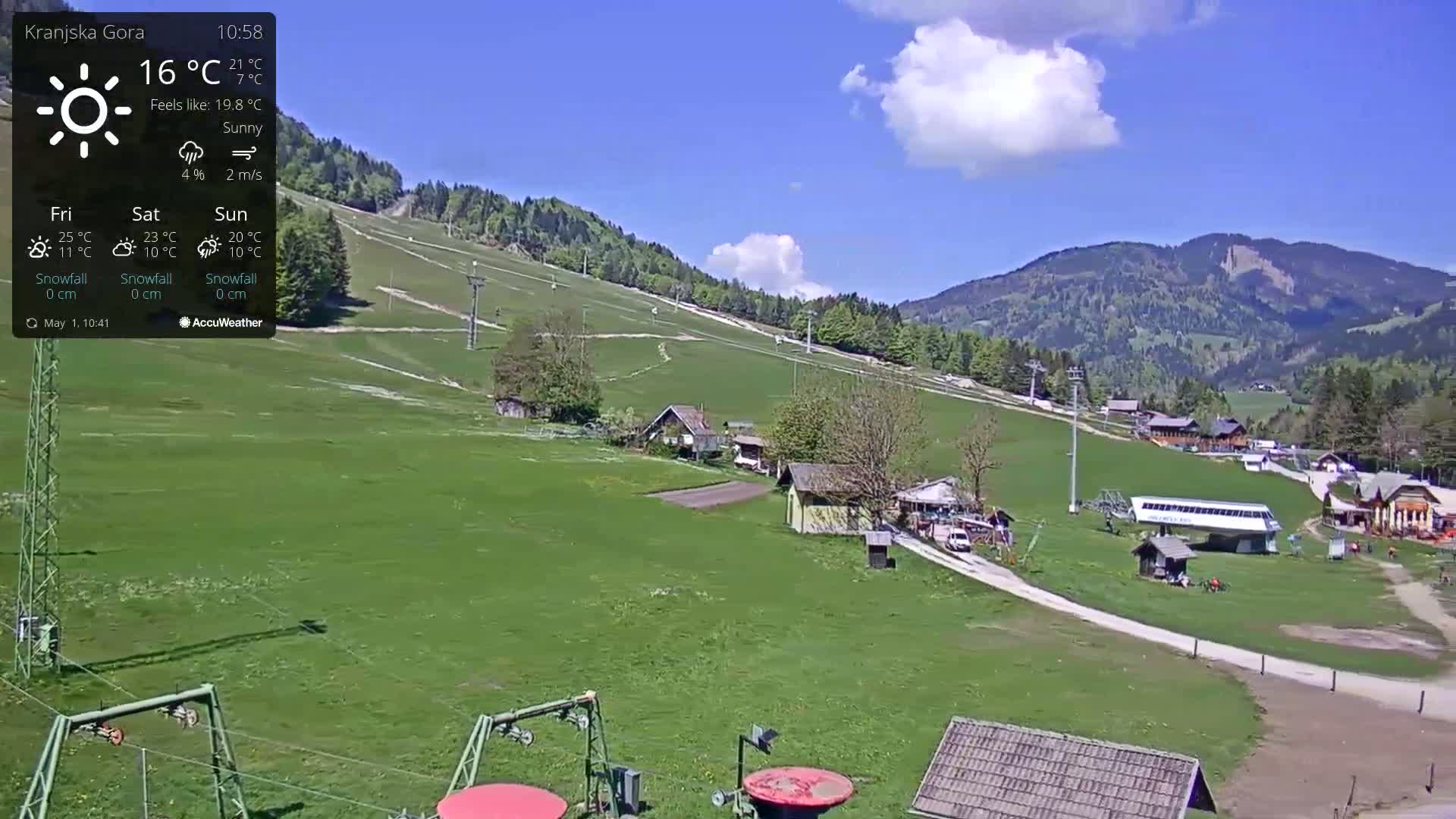 A sunny view of a grassy ski slope with several buildings and ski lifts, situated in a valley against a backdrop of mountains.