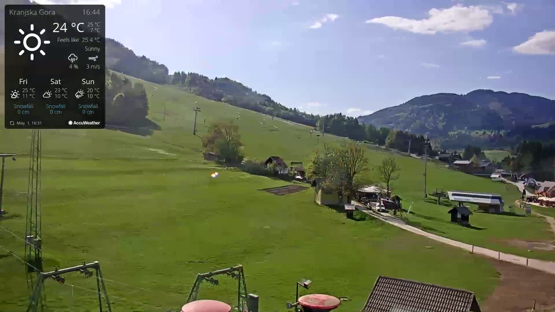 A sunny view of a grassy hillside with a few buildings and a chairlift, mountains in the background.