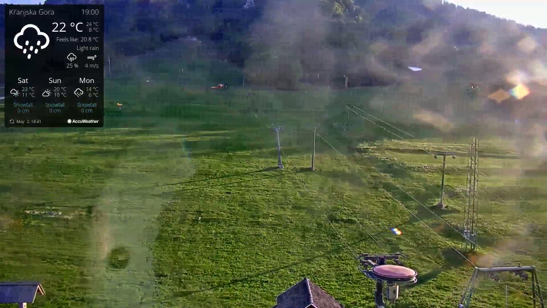 A high-angle view shows a grassy hillside with a chairlift and light rain under hazy sunlight.