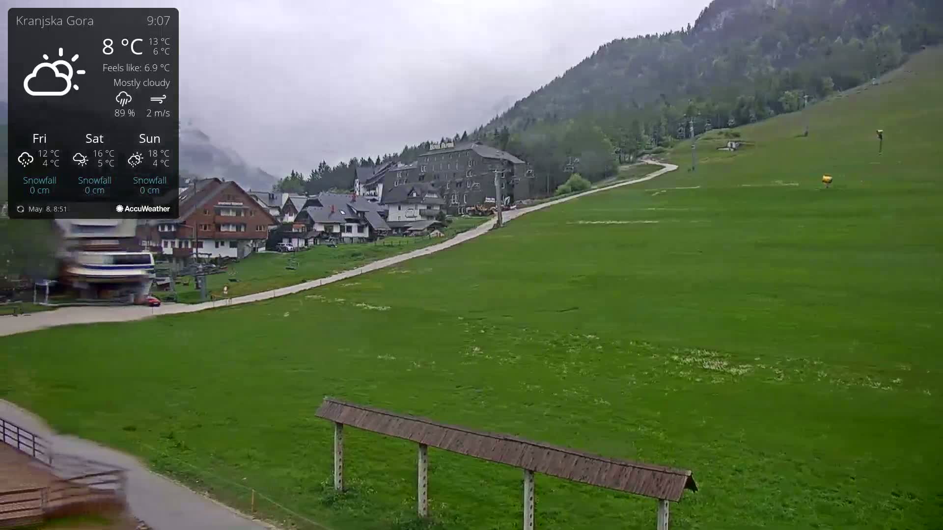 A mostly cloudy, slightly breezy day reveals a village nestled at the base of a mountain, with a grassy field and ski lift infrastructure in the foreground.