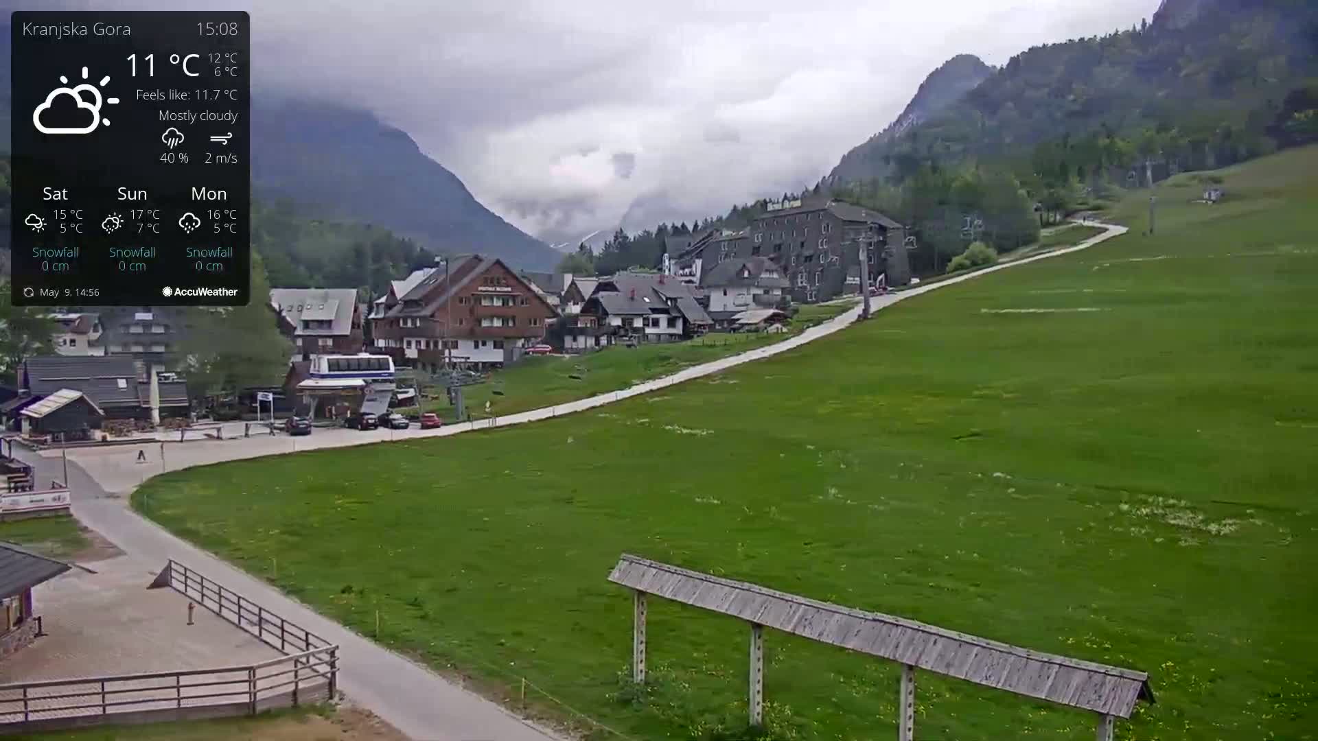 A mostly cloudy mountain village scene shows a paved road curving through a grassy field towards buildings and ski lifts, under overcast skies.