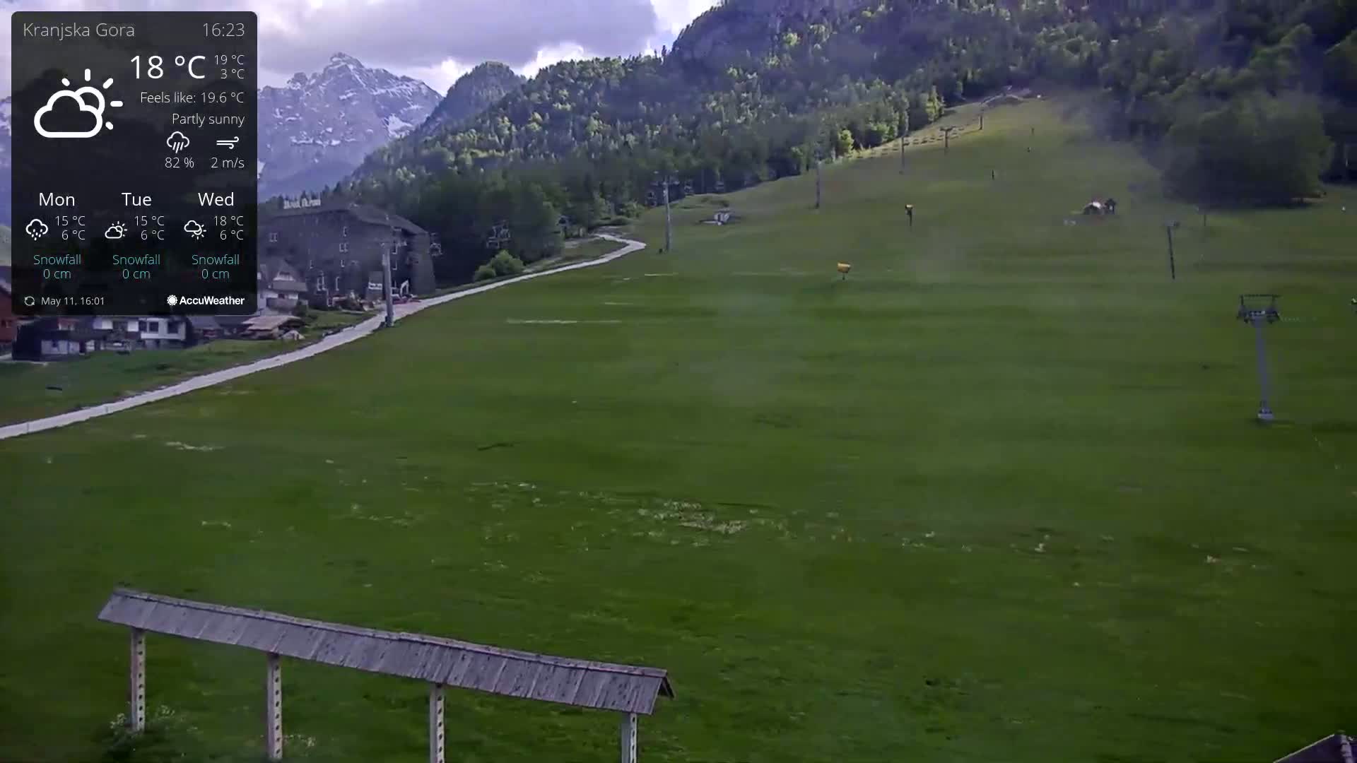 A partly sunny, green grassy slope with a ski lift and buildings in the background, nestled against mountainous terrain.