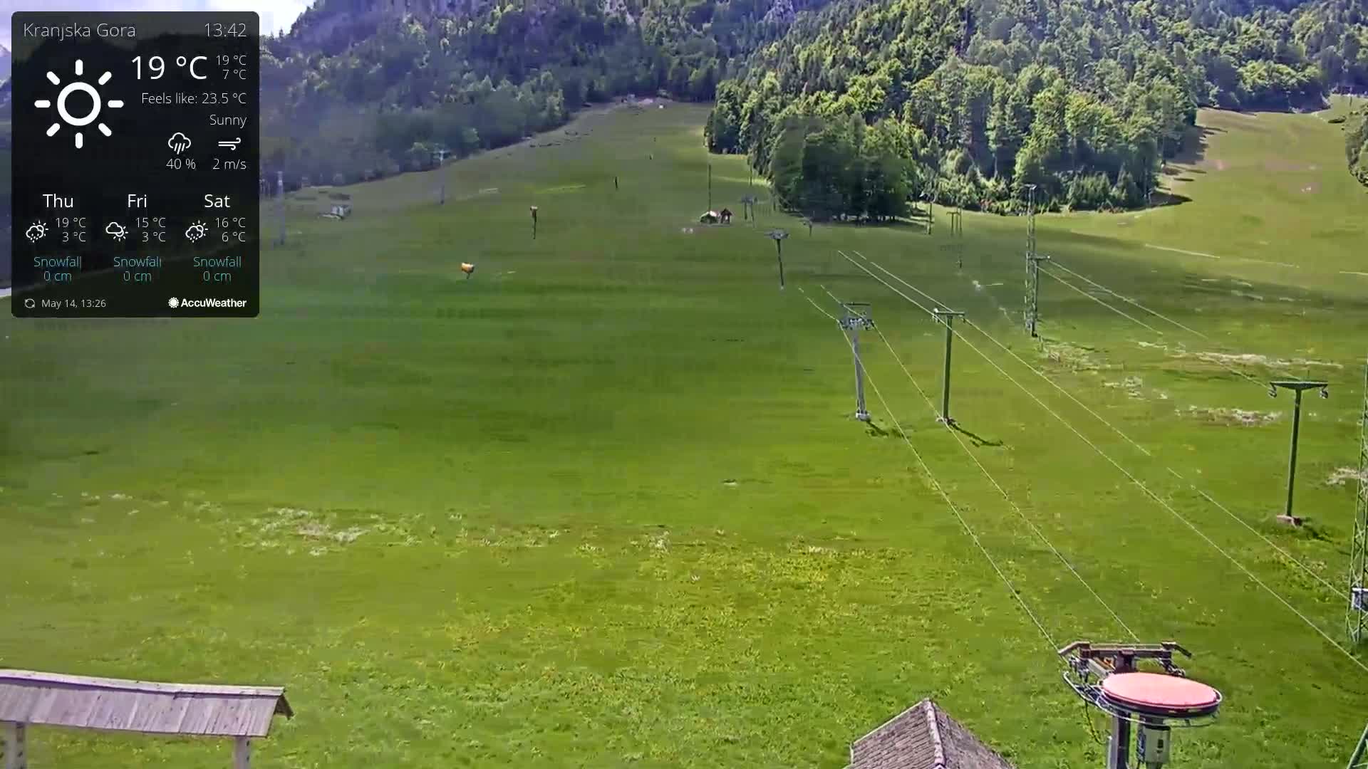 A sunny, high-angle view of a grassy field with a chairlift cable system and a few structures, including a small building and a covered shelter, in a mountainous region.