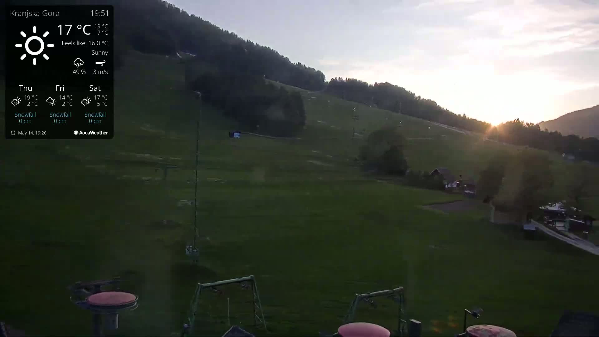 A sunny, but somewhat cool, sunset illuminates a grassy hillside with ski lifts and a few buildings,  some playground equipment visible in the foreground.