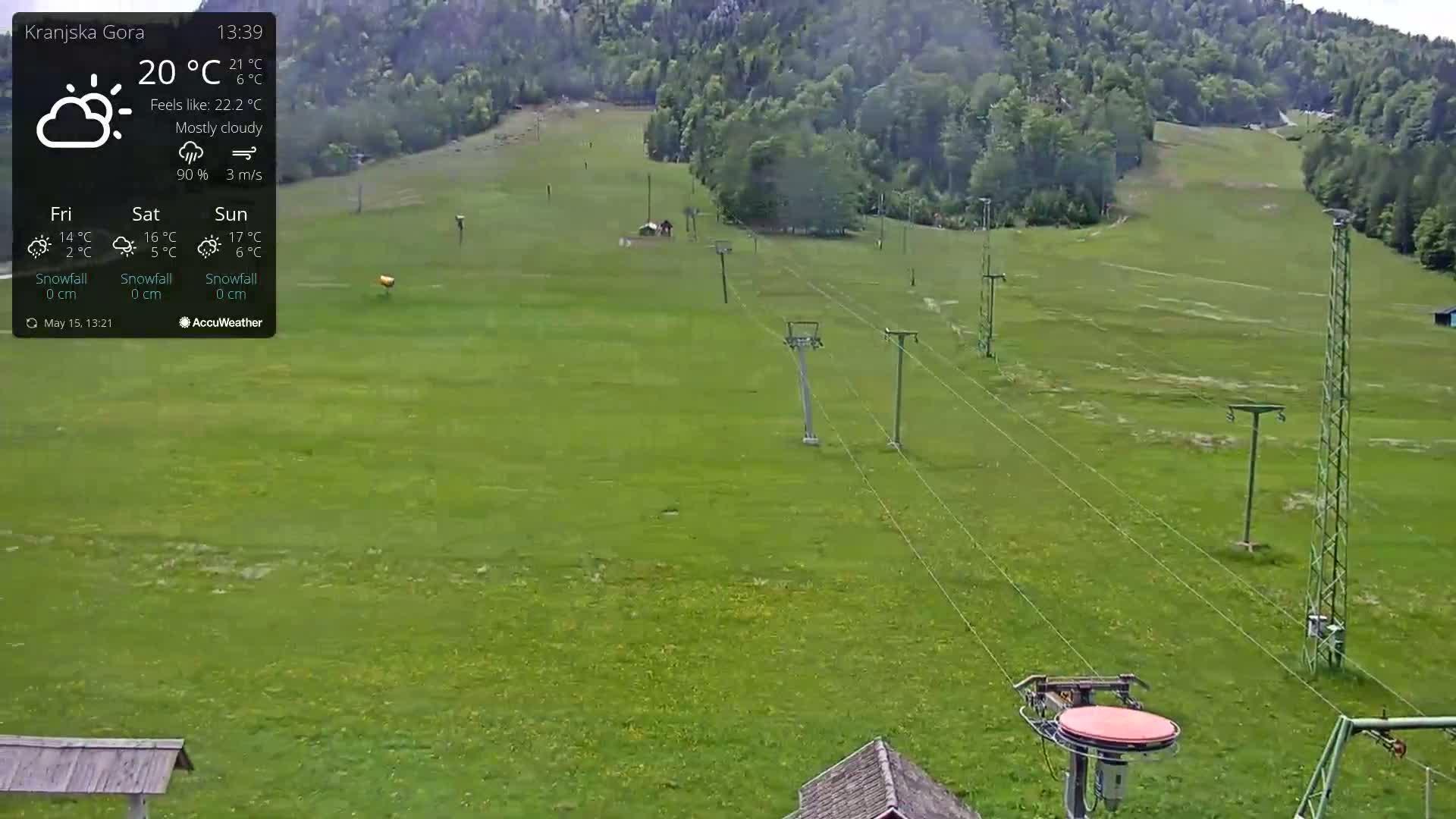 A mostly cloudy, green grassy field with a chairlift and tall metal poles stretches towards a tree-covered hillside.